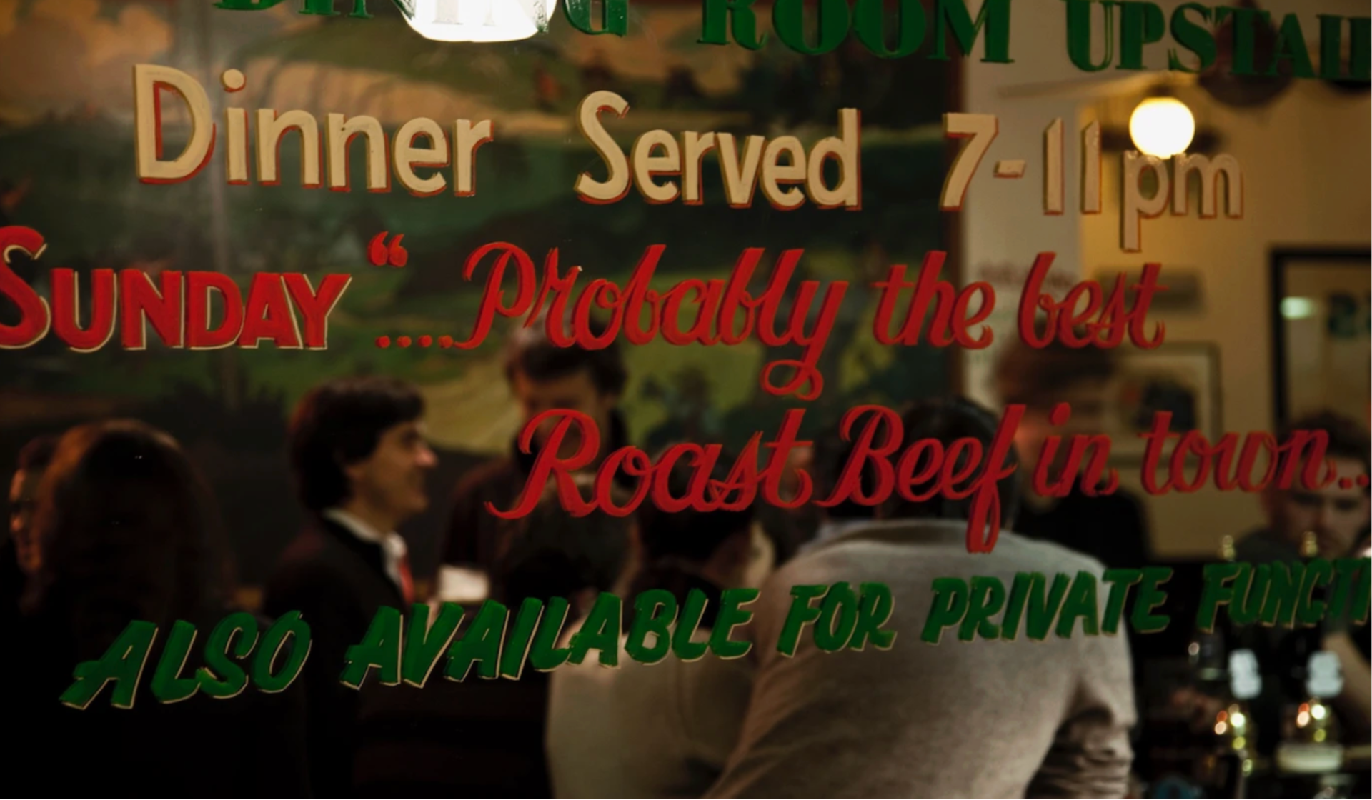 A glass window with painted text advertising dinner and roast beef reflects several people socialising inside a warmly lit restaurant, known for serving the best Sunday roast in London.
