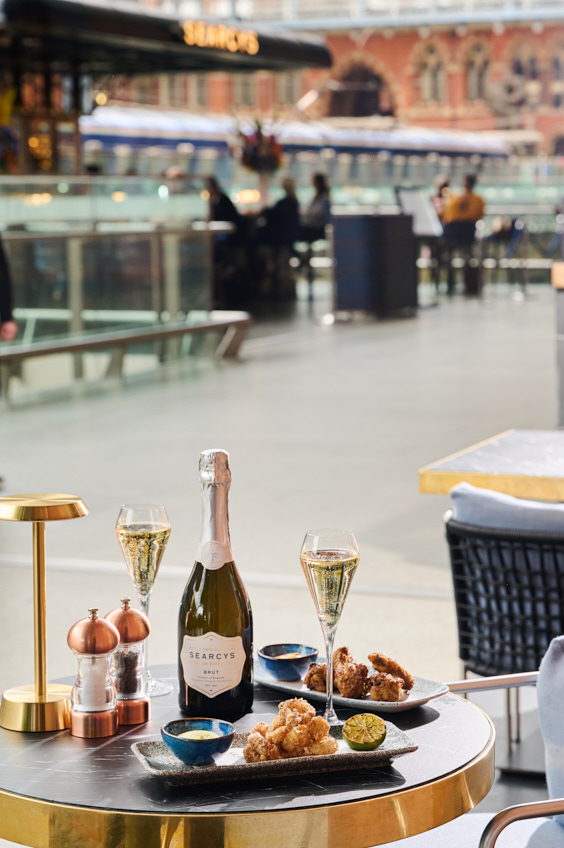 A table set with a bottle of Searcys sparkling wine, two glasses of bubbly, fried food, and condiments sits by a railing inside a busy, modern railway station—an ideal spot for your London weekend guide this December.