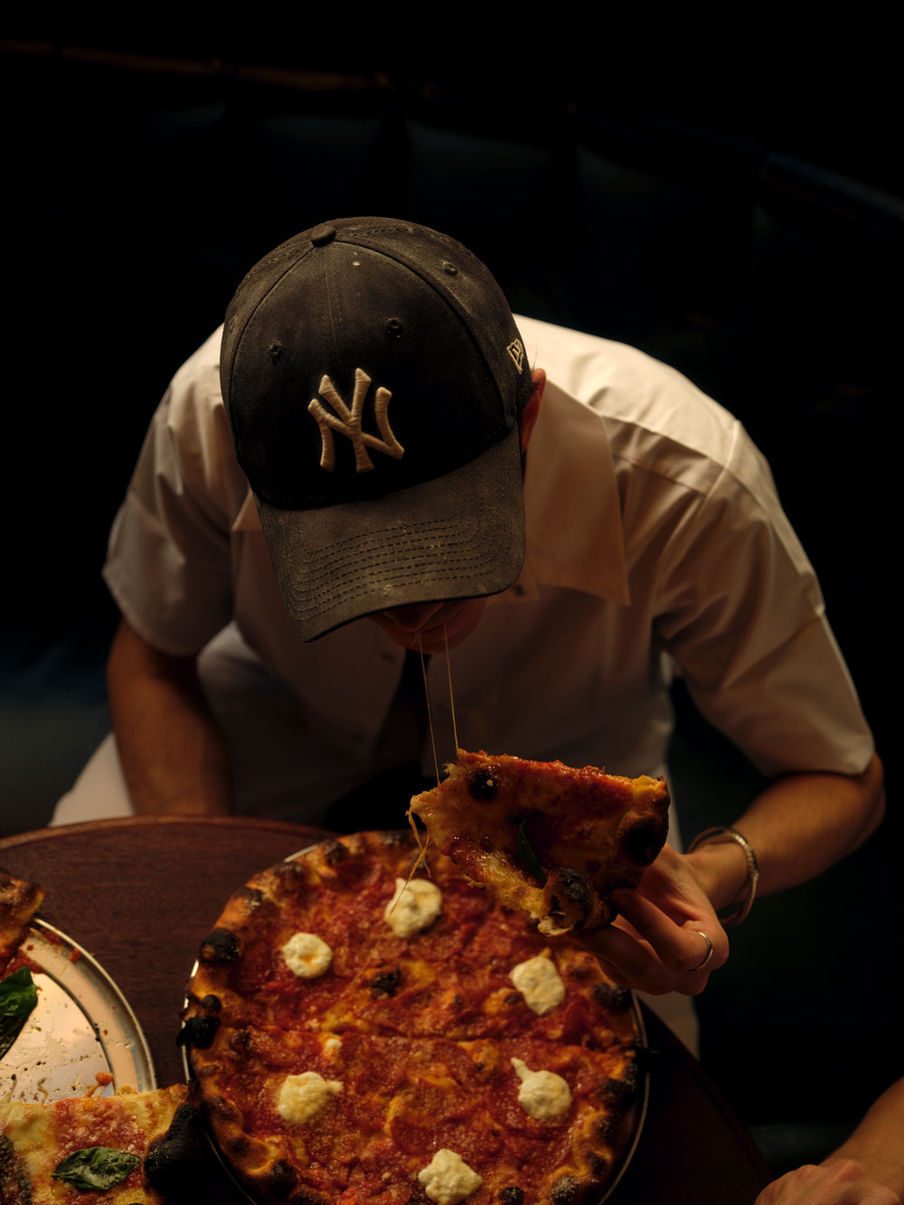 A person wearing a New York Yankees cap and white shirt eats a slice of pizza at a table, pulling up a cheesy slice from a pizza topped with tomato sauce and dollops of cheese—perhaps setting new pizza standards for London’s pizza scene.