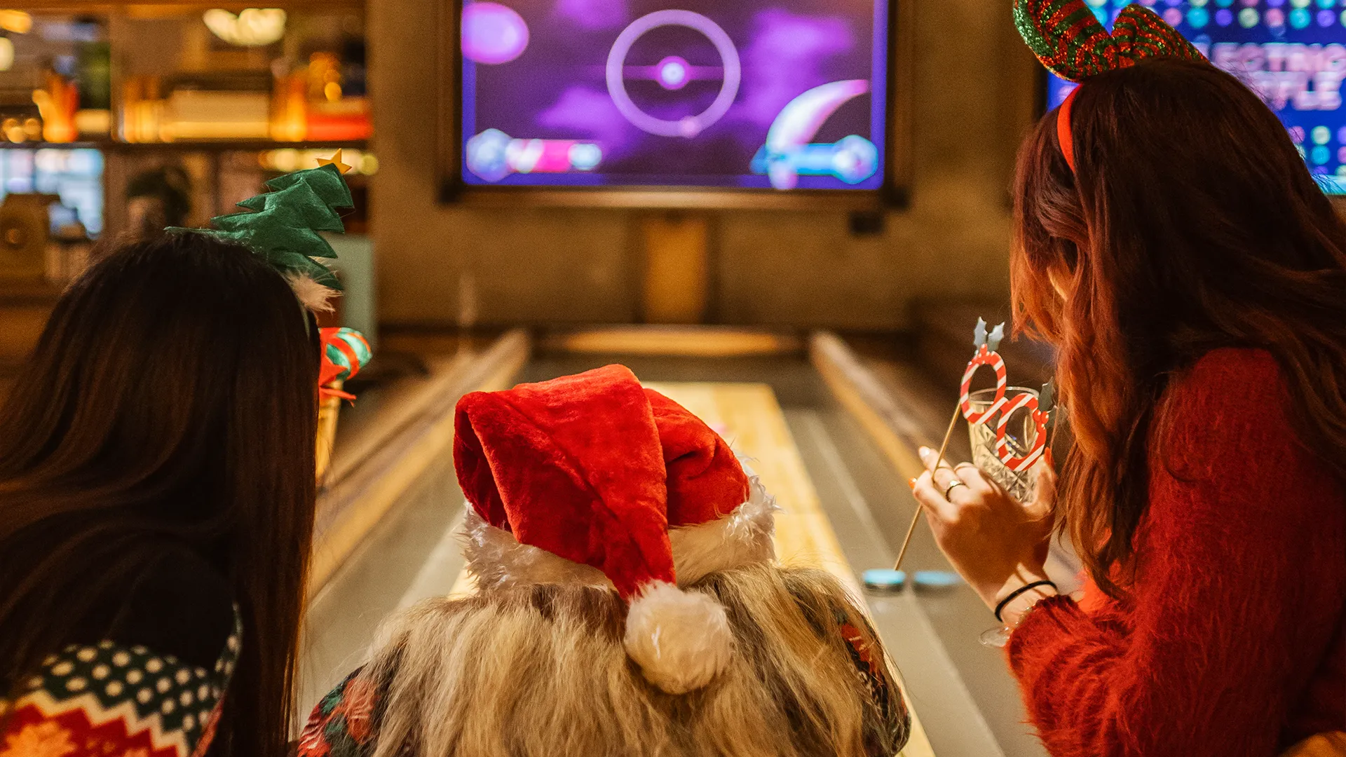 Three people in festive holiday hats enjoy a lively bowling alley, watching the lane as a large screen glows in the background—a perfect scene for your London weekend guide this December.