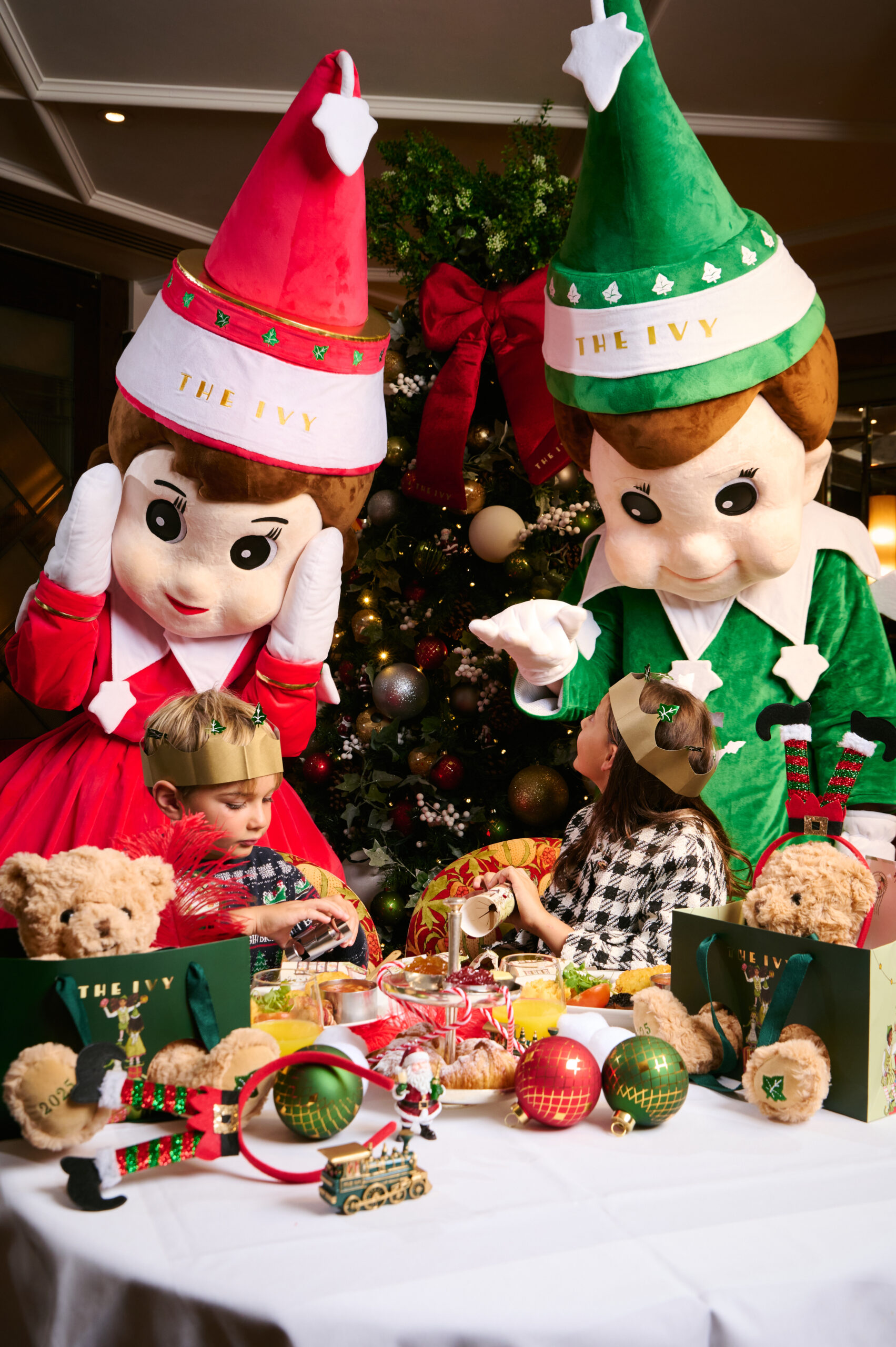 Two children in paper crowns sit at a festive table with Christmas treats and teddy bears, while two people in large elf costumes pose behind them—a perfect scene for your London weekend guide December. A decorated Christmas tree sparkles in the background.