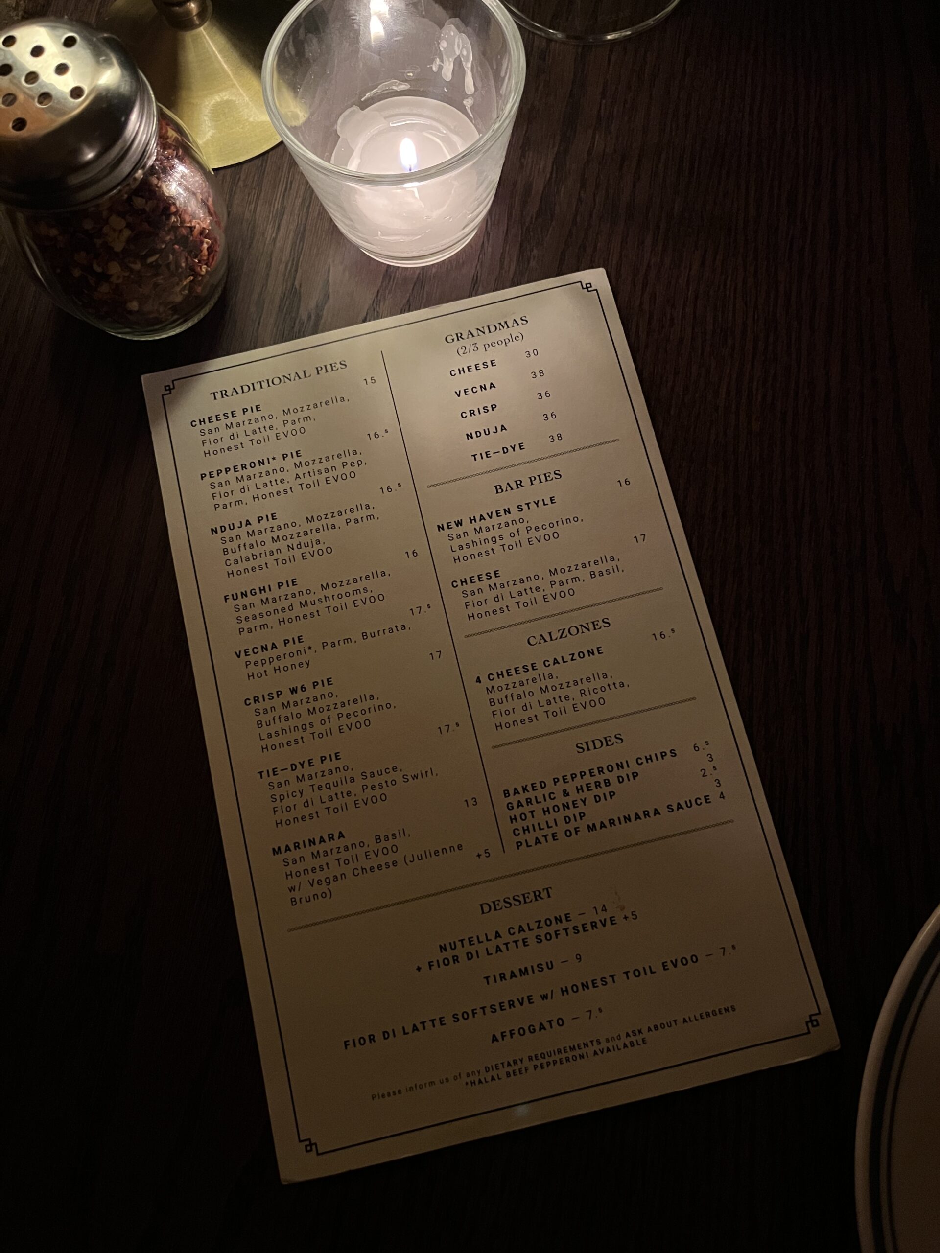 A restaurant menu on a dark wooden table is lit by a candle. The menu lists traditional pies, grandma pies, bar pies, calzones, sides, and desserts—including signature London pizza. A pepper shaker and glass are also visible in the background.