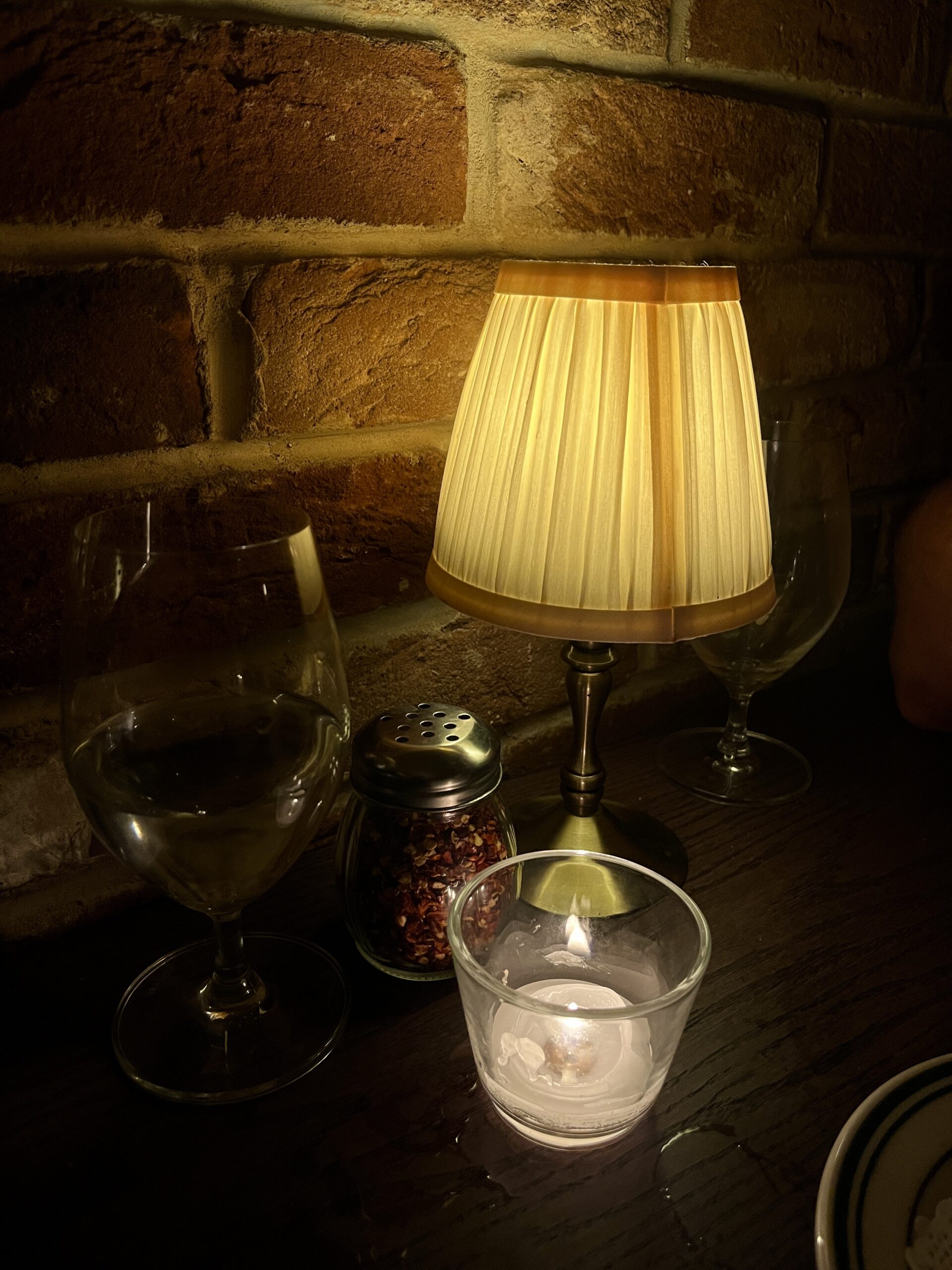 A cosy table setting with a small lamp, two empty wine glasses, a glass candle holder with a lit candle, a spice shaker, and a brick wall background evokes the charm of London's pizza standards under warm yellow light for a relaxed atmosphere.