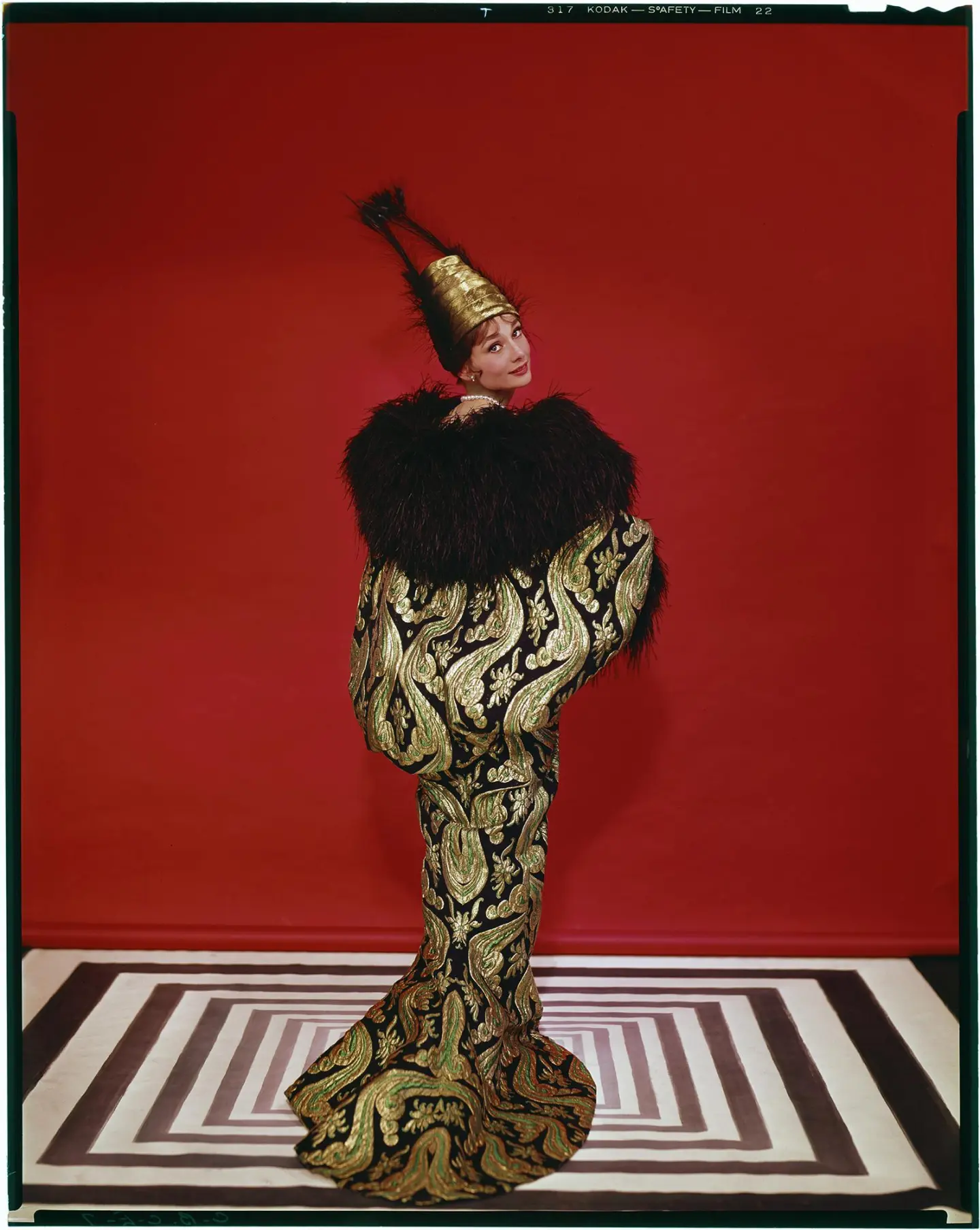 A person poses in front of a red backdrop, wearing a gold and black patterned gown with a large feathered collar and a tall, gold conical hat—an eye-catching look perfect for the London weekend guide December edition. They stand on a black-and-white geometric floor.