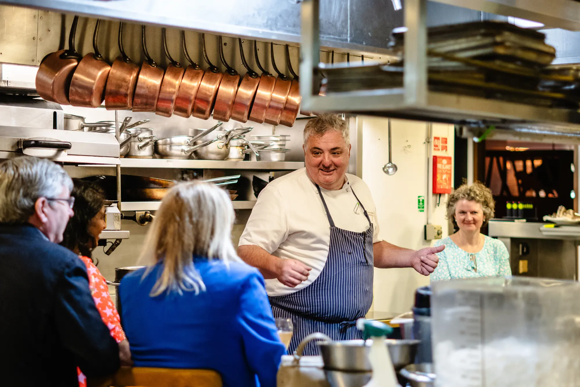 A chef in a striped apron talks and smiles with a group of seated people in a professional kitchen—a cosy scene perfect for any London weekend guide December edition, with pots, pans, and cooking equipment adding to the festive atmosphere.