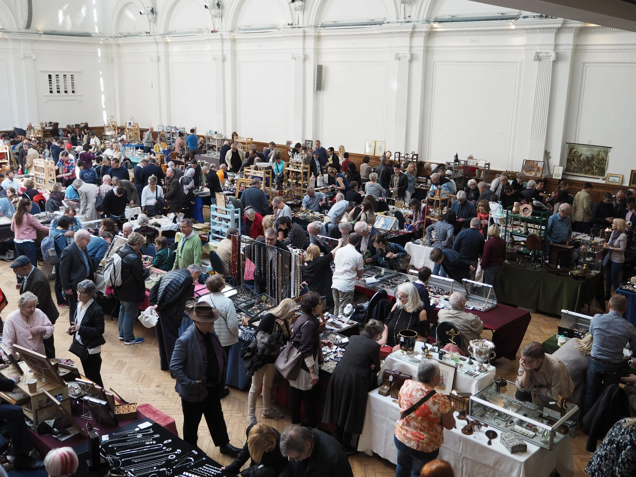 A large group of people browse and shop at various vendor tables displaying antiques and collectables inside a spacious, well-lit hall—a highlight in any London weekend guide for December.