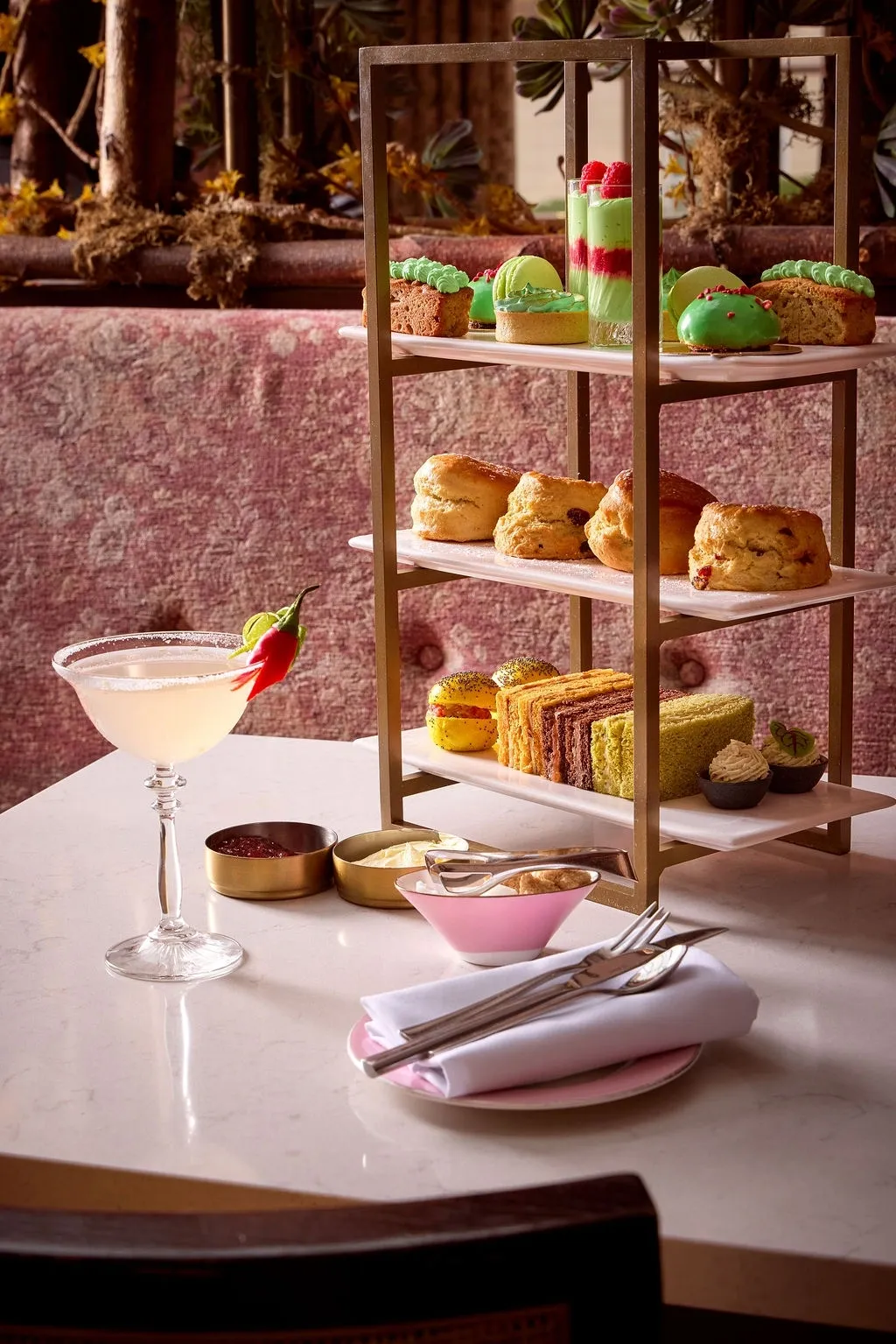 A three-tier afternoon tea stand holds various pastries, scones, macarons, and finger sandwiches—perfect for a festive treat in your London weekend guide December. A chilli-garnished cocktail sits beside jam, cream, plate, serviette, and cutlery on the table.