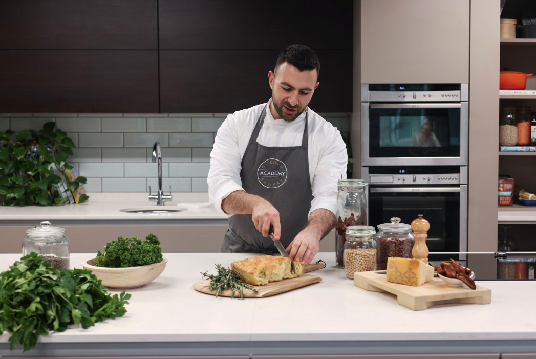 A chef in a modern kitchen slices bread on a wooden board, surrounded by fresh herbs, grains, and cheese—an inviting scene that could inspire any London weekend guide December food adventure. Shelves, ovens, and a sink complete the cosy backdrop.