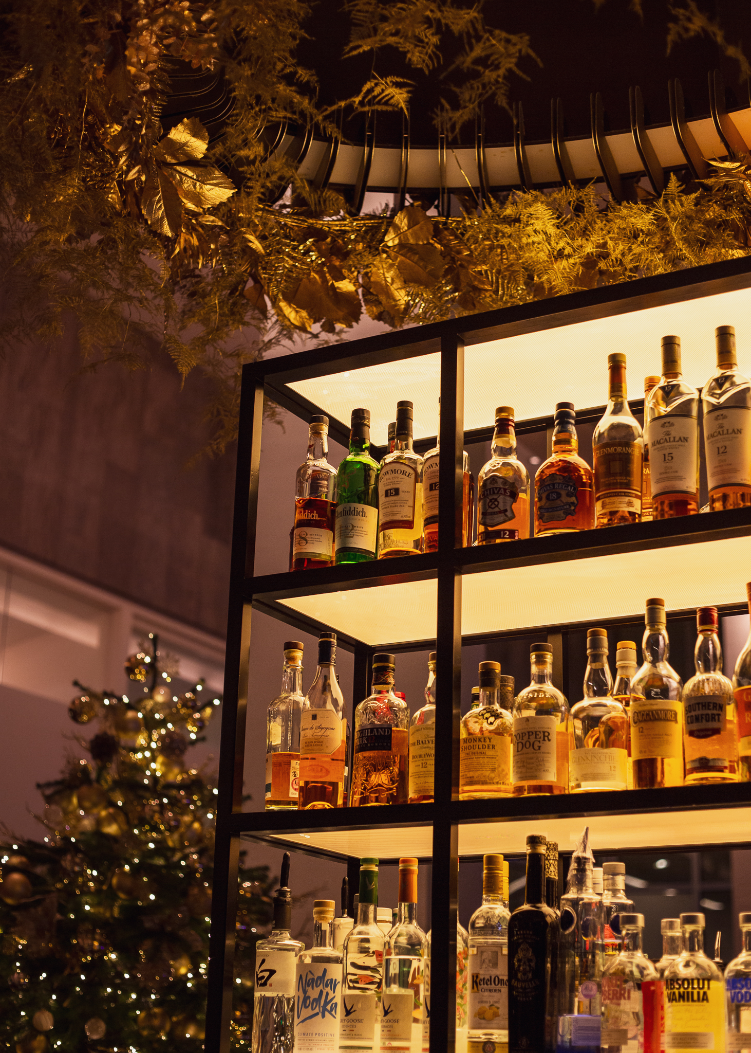 A backlit bar shelf displays various bottles of liquor, with golden decorative leaves above and a lit Christmas tree in the blurred background, creating a festive atmosphere perfect for any London weekend guide December edition.