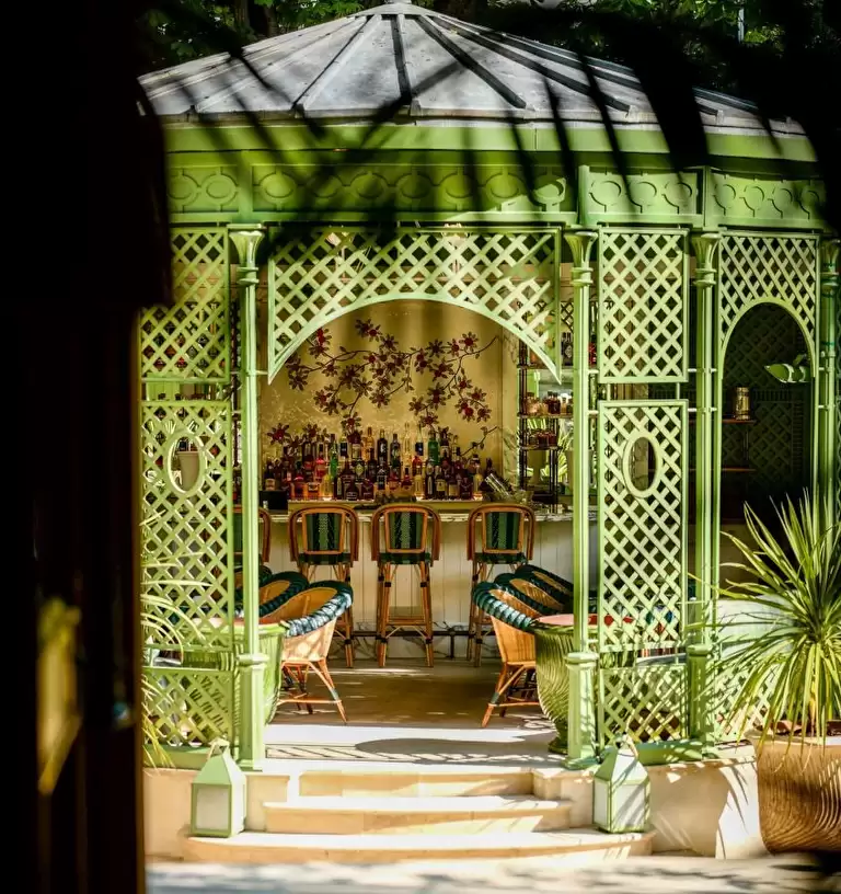 A green gazebo with lattice walls shelters a cosy bar, featuring wicker chairs and a counter lined with bottles—perfect for unwinding after a train journey from London amid sunlight and palm shadows that create a relaxed, tropical atmosphere.