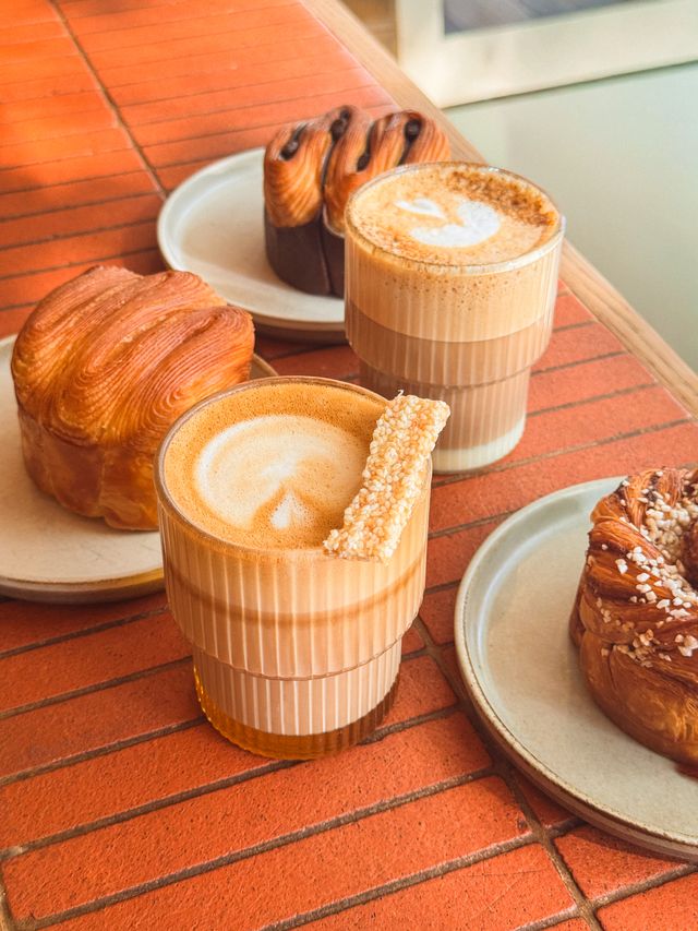 Two lattes in ribbed glasses, each on a plate with golden, flaky pastries, are arranged on a red brick table—an inviting scene for your weekend guide to cosy café indulgence. One latte is topped with a playful pastry stick garnish.