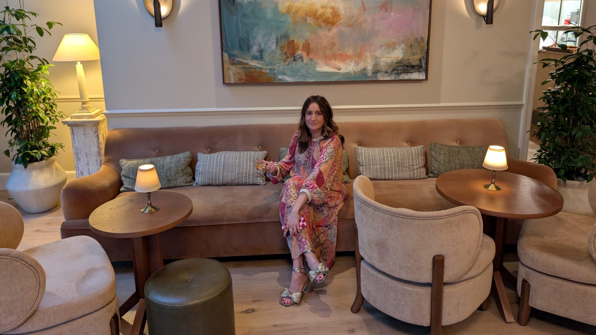 A fashion editor in a colourful dress sits on a beige sofa in a stylish, modern living room with soft lighting, abstract wall art, two armchairs, side tables with lamps, and green plants on either side.