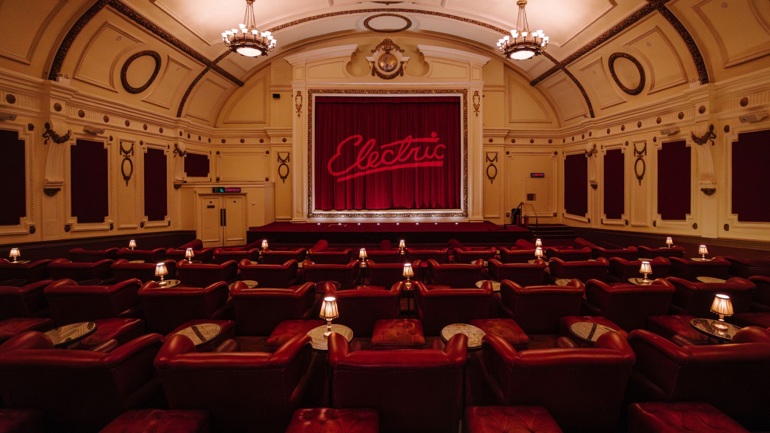 A vintage-style cinema, among the finest London independent cinemas, with plush red armchairs, small round tables, and lamps. The stage has a red curtain with Electric written on it, and ornate chandeliers hang from the curved decorative ceiling.