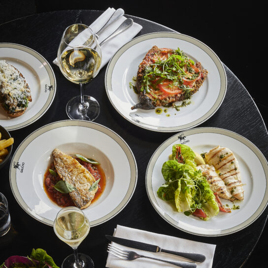 A round black table set for a refined meal, perfect for your weekend guide, features four plates of gourmet food—fish, fresh salad, and vibrant garnished dishes—alongside a glass of white wine, champagne, cutlery, and a folded serviette.