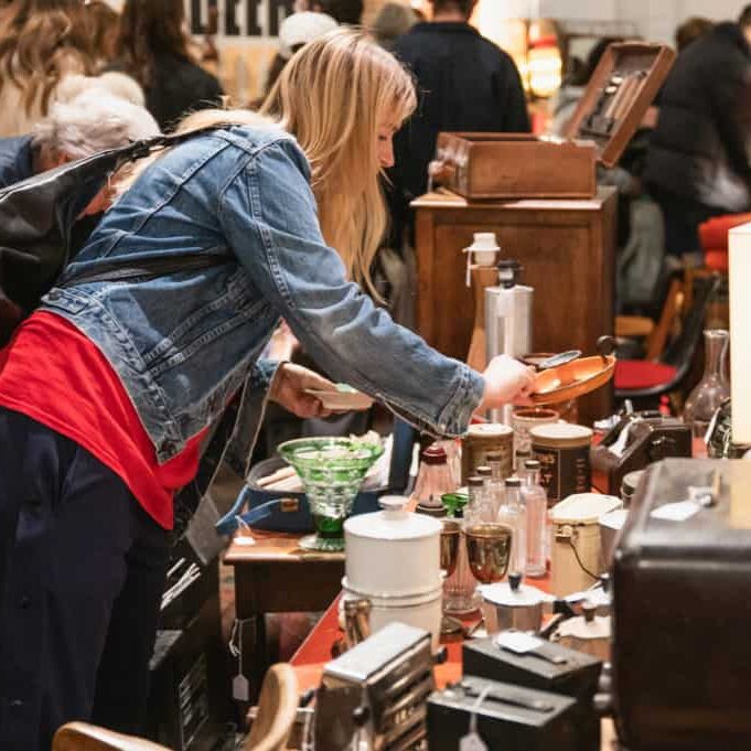A woman in a denim jacket browses vintage items on a crowded table at an indoor London flea market, surrounded by other shoppers and eclectic antiques.