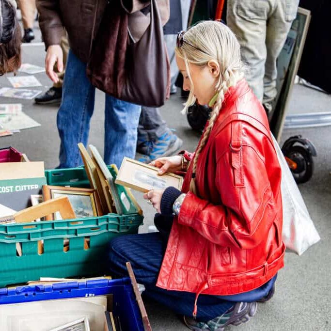 A person in a bright red jacket kneels on the ground, looking through framed pictures in a green crate at what appears to be one of the vibrant London flea markets, with other people browsing nearby.