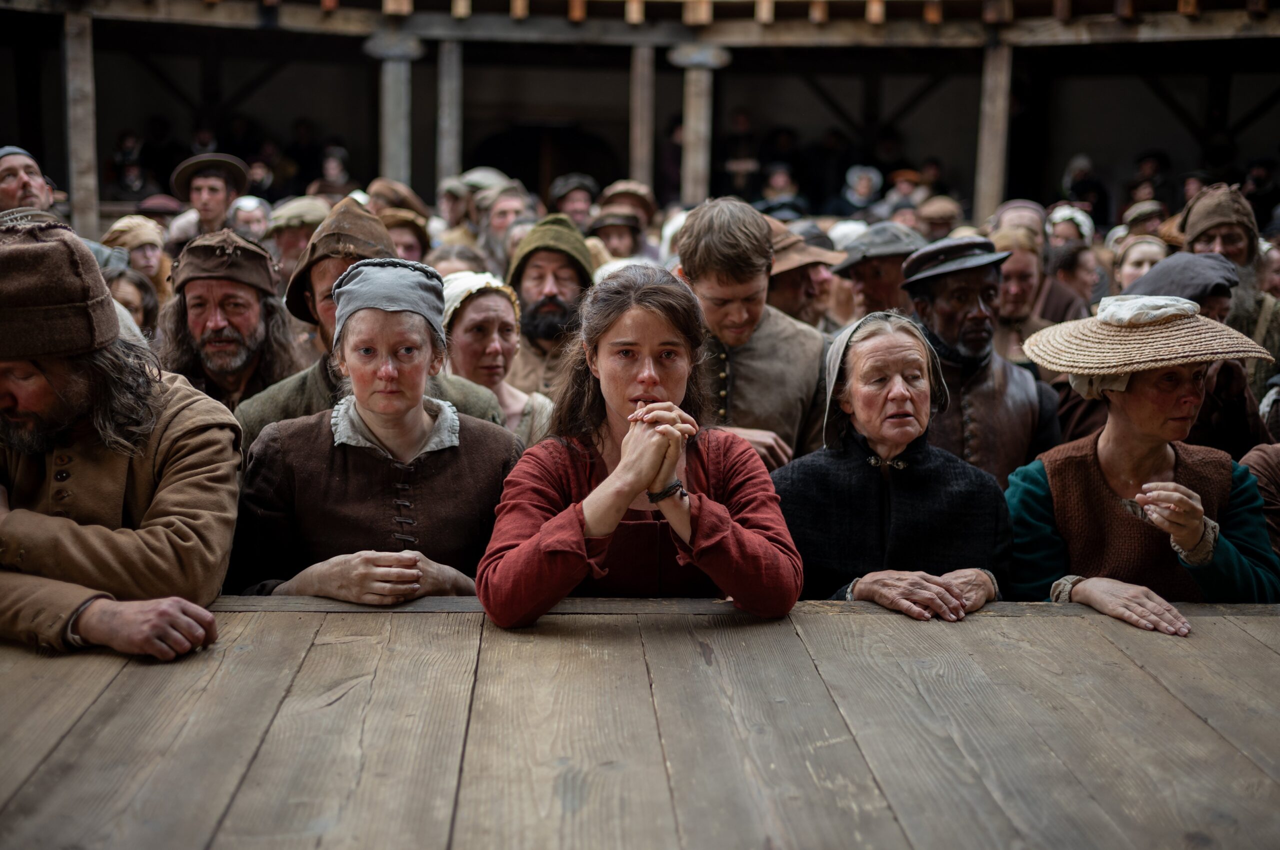 A group of people in old-fashioned, rustic clothing stand closely together in front of a wooden barrier, looking serious and sombre, as if attending a historical event or trial from the time of Hamnet.
