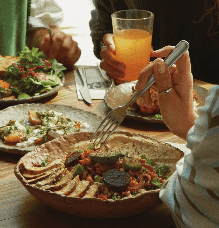 Three people enjoy a vibrant meal at a wooden table during London wellness days, savouring salad, roasted vegetables, and orange juice. One holds a fork above their food, while another lifts a glass of juice.