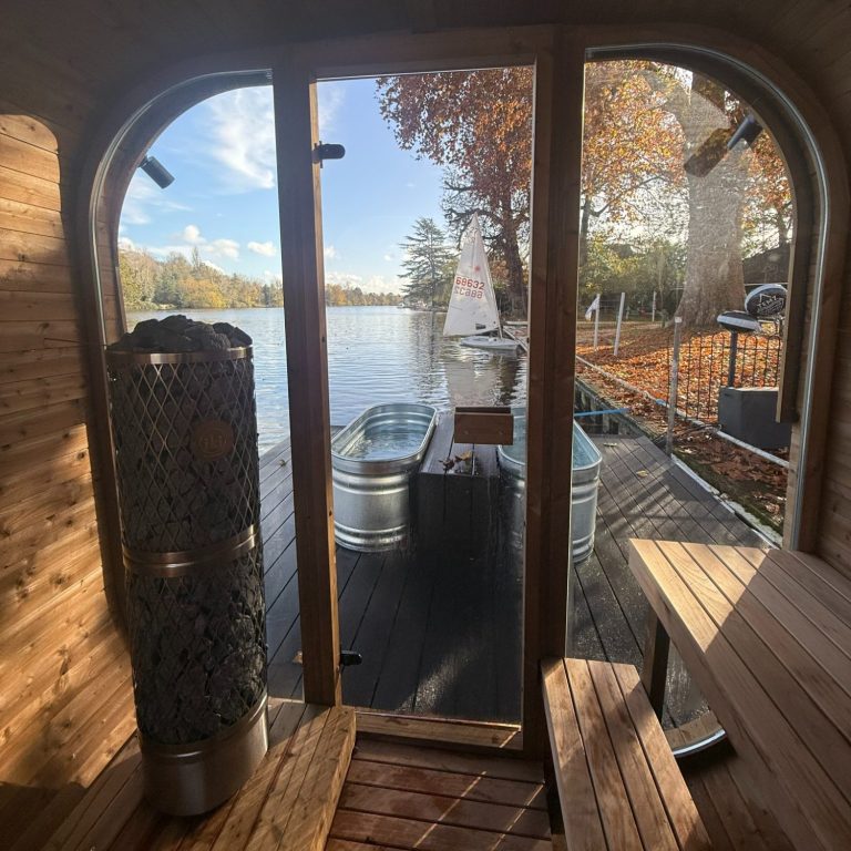 View from inside a wooden sauna, looking out through a glass door onto a lakeside deck with metal tubs and autumn trees. Sunlight streams in, creating a warm, inviting scene—perfect inspiration for relaxing London wellness days.