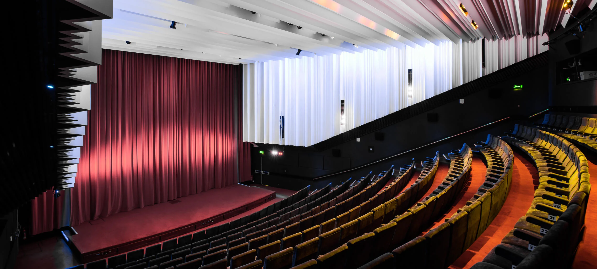 Empty modern cinema auditorium with colourful tiered seating, angled rows, a red carpeted stage, and a large red curtain covering the screen—reminiscent of stylish London independent cinemas. White and black panelled walls and ceiling complete the look.