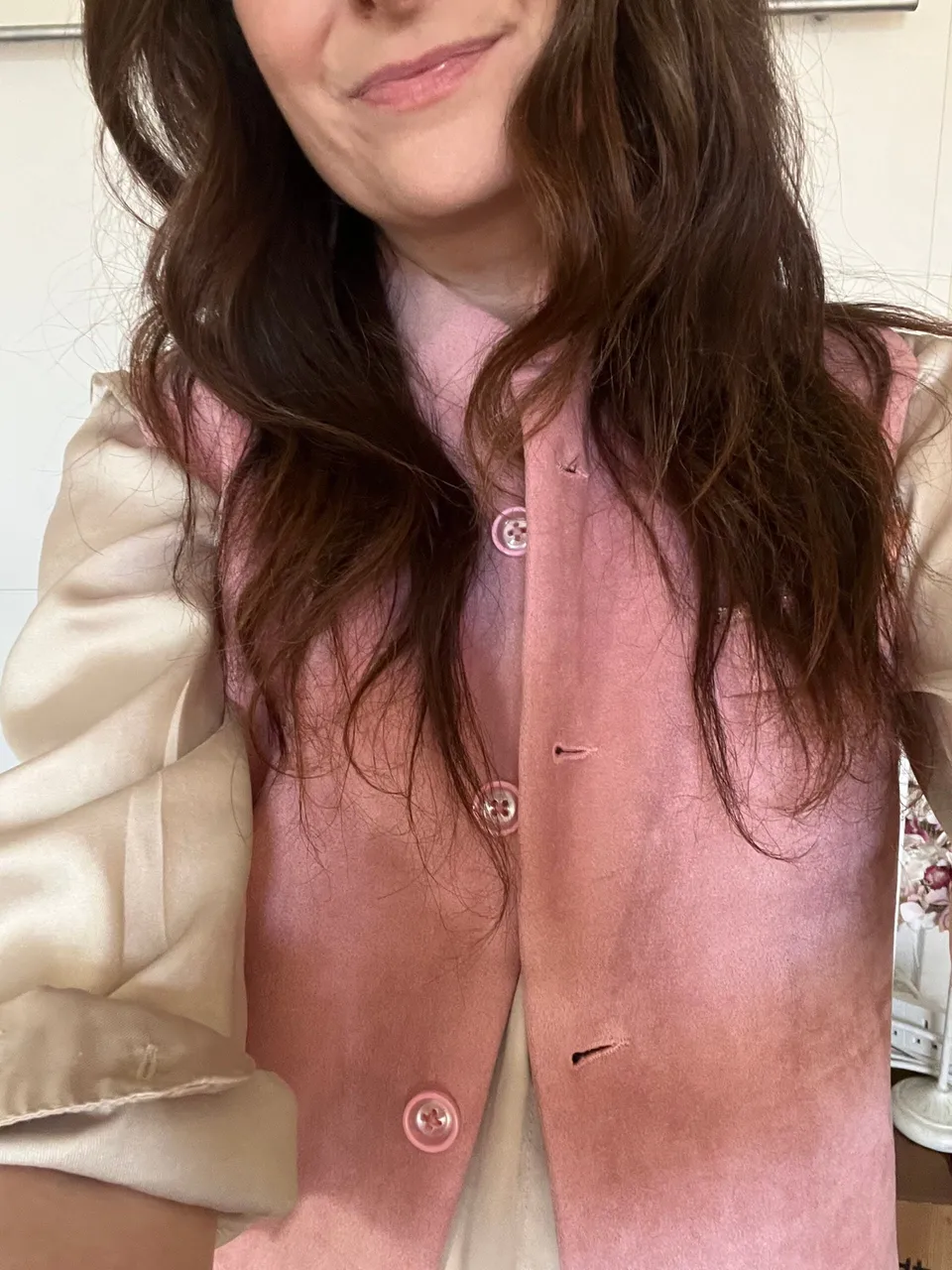 A woman with wavy brown hair, reminiscent of a Fashion Editor, wears a pale pink waistcoat over a cream-coloured blouse. She is smiling slightly and only her upper body is visible in the image.