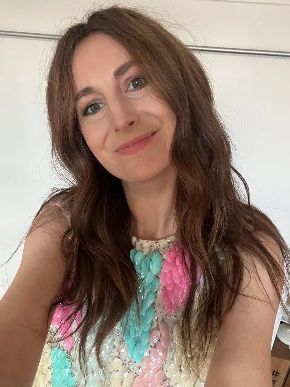 A woman with long, wavy brown hair smiles at the camera, embodying life in fashion. She is wearing a sleeveless top with pastel blue, pink, and green embellishments, standing in front of a plain white background.