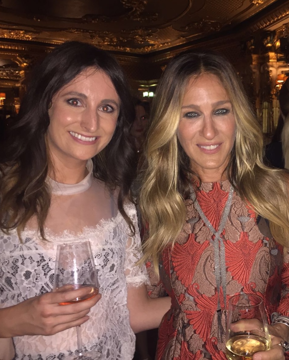 Two women smiling and posing together at an indoor event, each holding a glass of wine. The ornate gold decor and warm lighting create an elegant, festive atmosphere—perfect for anyone who loves Life In Fashion.