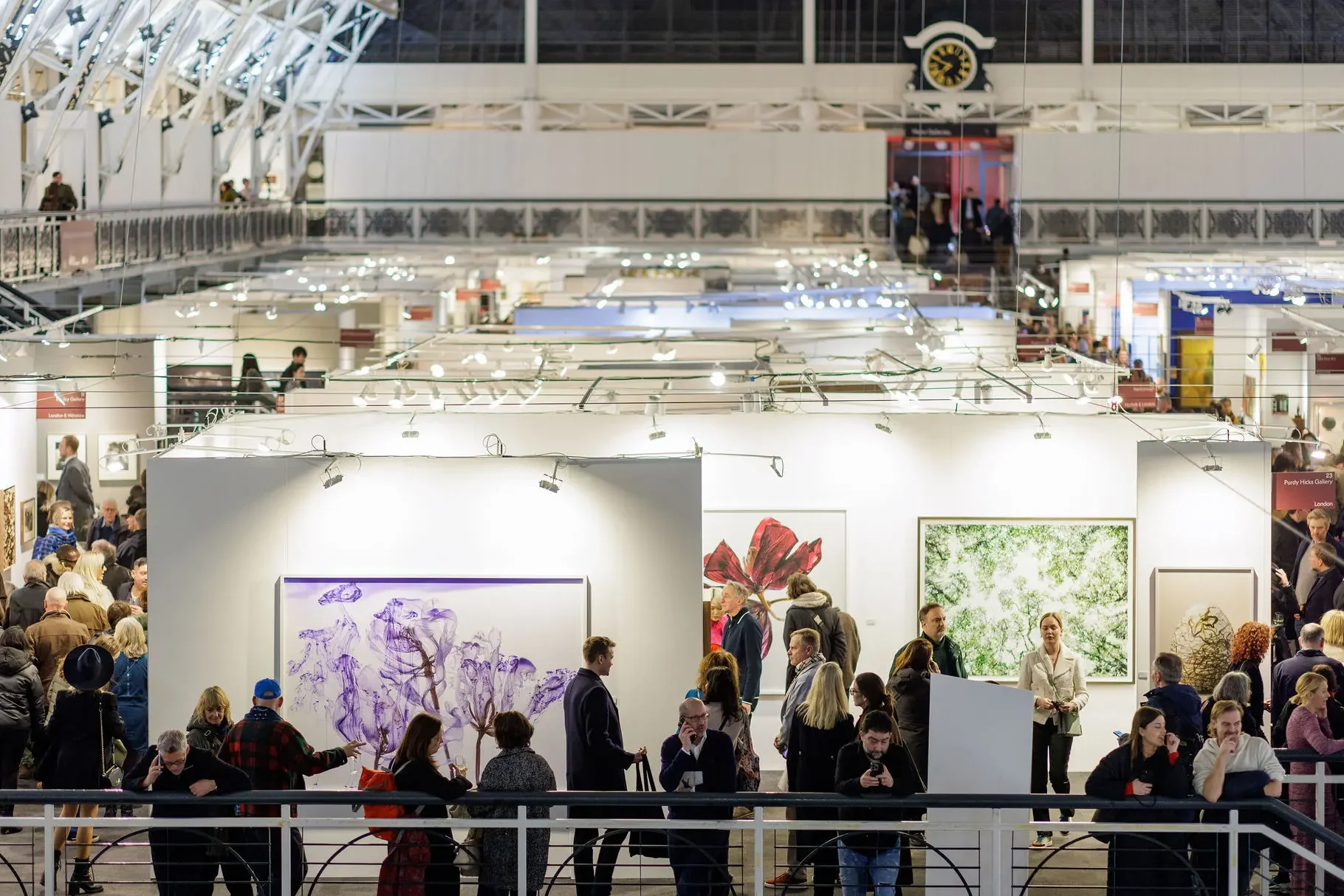 A crowded indoor art exhibition, perfect for your London weekend guide, features people viewing and discussing large artworks displayed on white walls under bright lights, beneath a high arched ceiling with a prominent clock in the background.