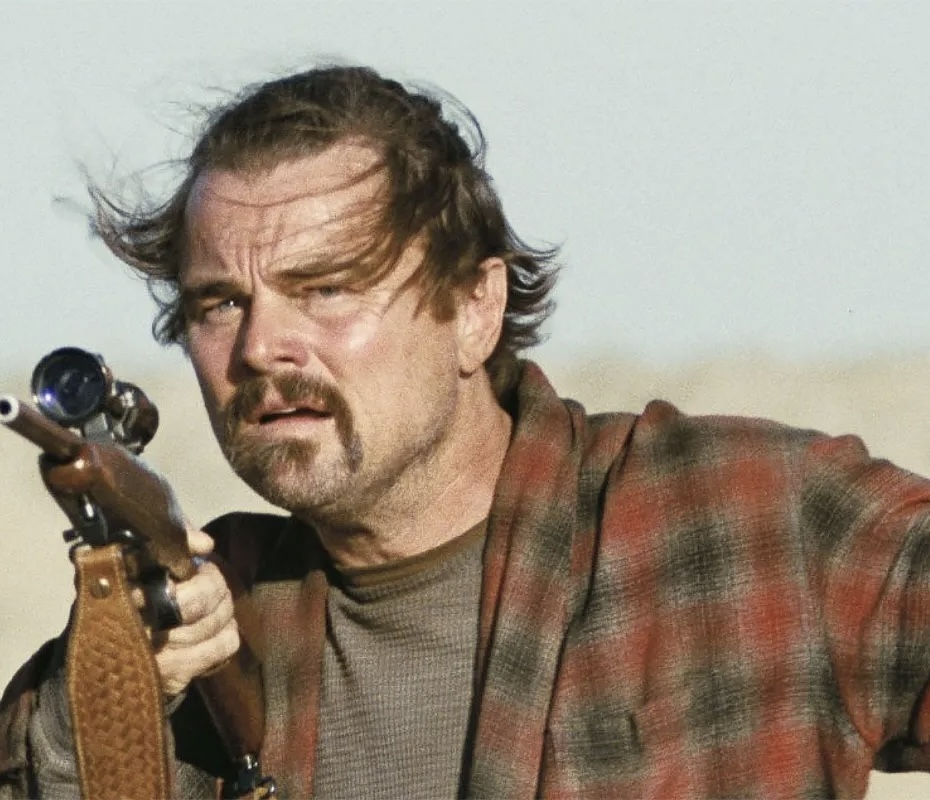 A man with tousled hair and a goatee, like a character from awards season films, wears a checked jacket and holds a rifle, looking intently into the distance outdoors against a blurred background of sand or sky.