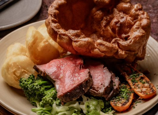 A plate of roast beef slices, Yorkshire pudding, roast potatoes, broccoli, cabbage, and carrots topped with herbs—a classic dish to enjoy on any London weekend guide.