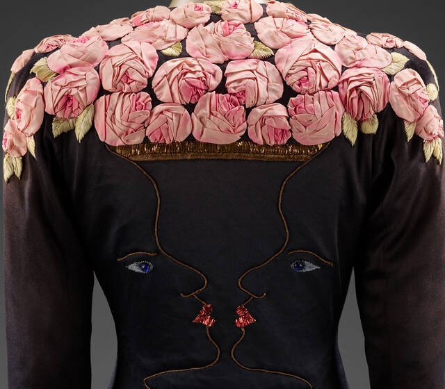 A black garment with embroidered faces in profile, sharing lips, forms a vase-like shape. Ideal for Fashion Exhibitions, its top is crowned with pink, three-dimensional fabric roses and gold leaves, creating an artistic, surreal effect.