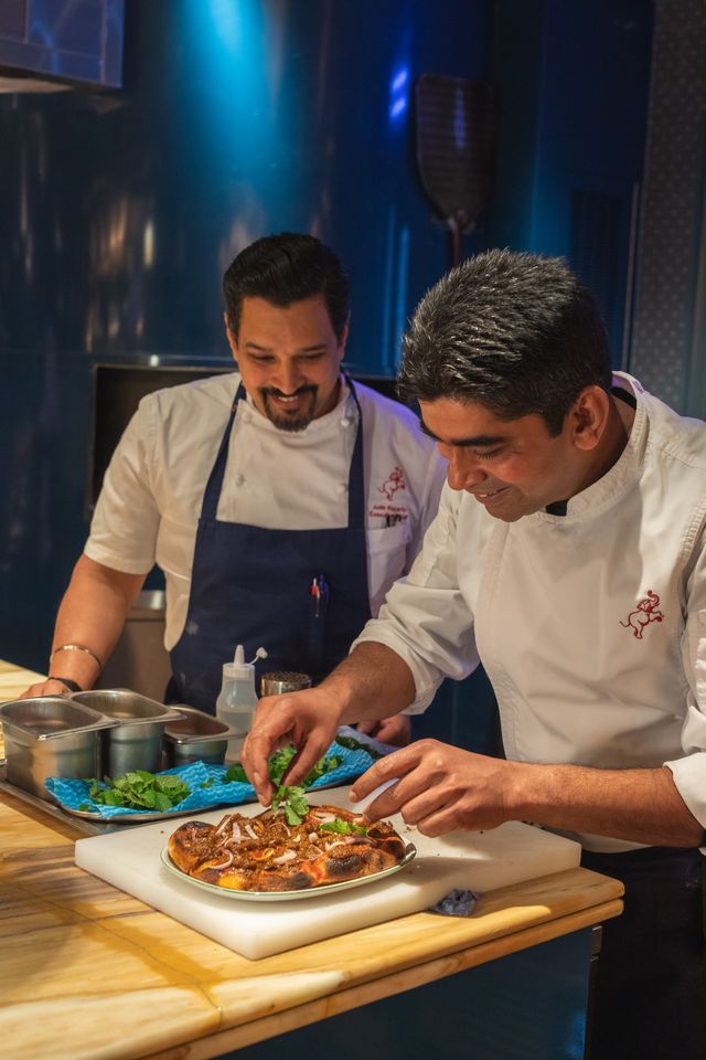 Two chefs in white uniforms, smiling and focused, prepare and garnish a dish together in a professional kitchen. Various tools and ingredients hint at special February pop-ups or delights from the Valentines line on the counter.