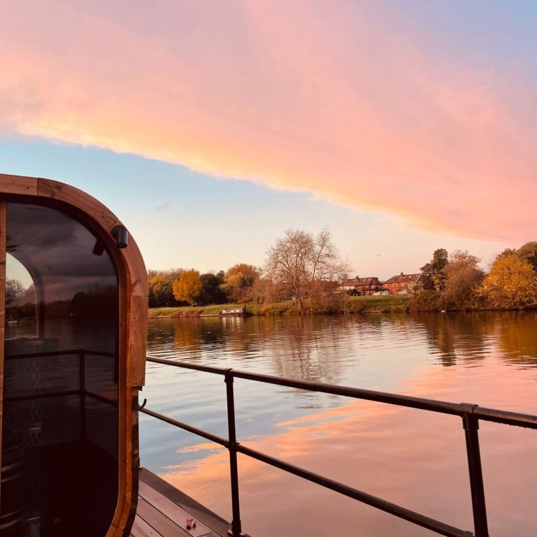 A riverside view at sunset with pink clouds reflecting on the calm water. Part of a wooden structure, possibly a houseboat, is visible to the left, evoking the peaceful retreat of London wellness days. Trees with autumn foliage line the far riverbank.