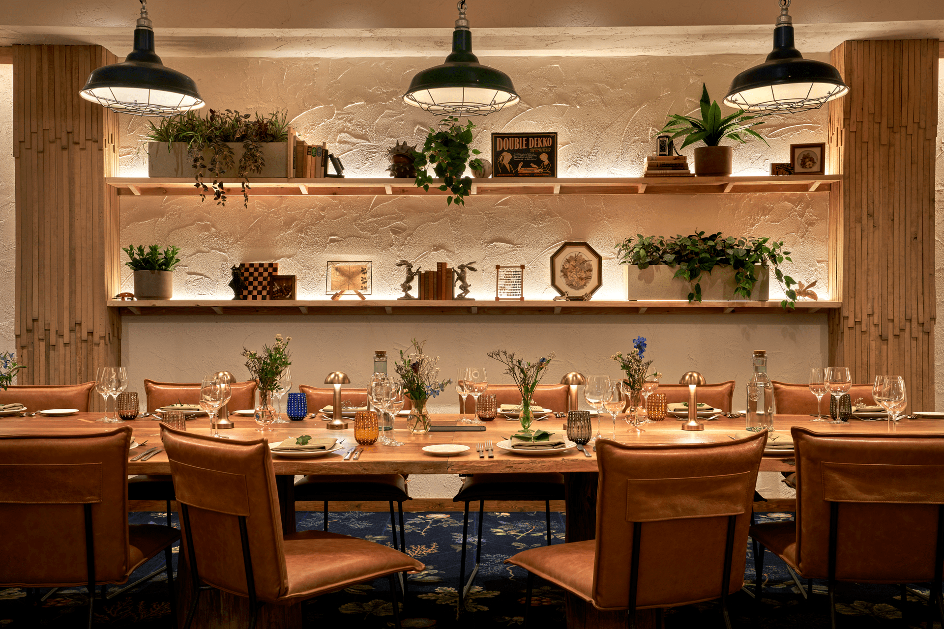 A warmly lit dining room with a long wooden table set for a meal, surrounded by leather chairs. Shelves on the back wall display plants, books, and decorative items under soft ambient lighting—perfect for a Manchester Guide feature.