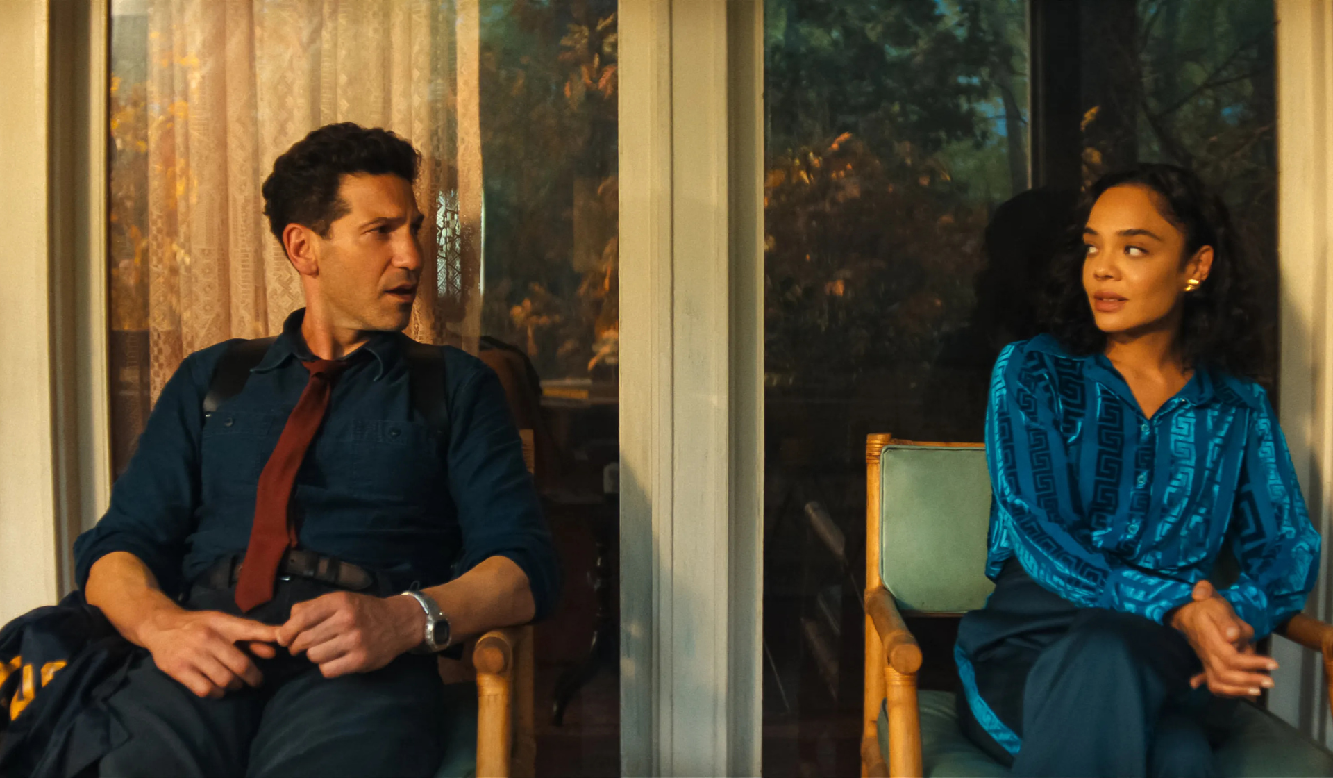 A man and a woman sit on chairs outside, facing each other like characters in a Netflix thriller. The man wears a dark shirt with a loosened red tie, while the woman wears a blue blouse and black trousers. A window and trees are visible in the background.