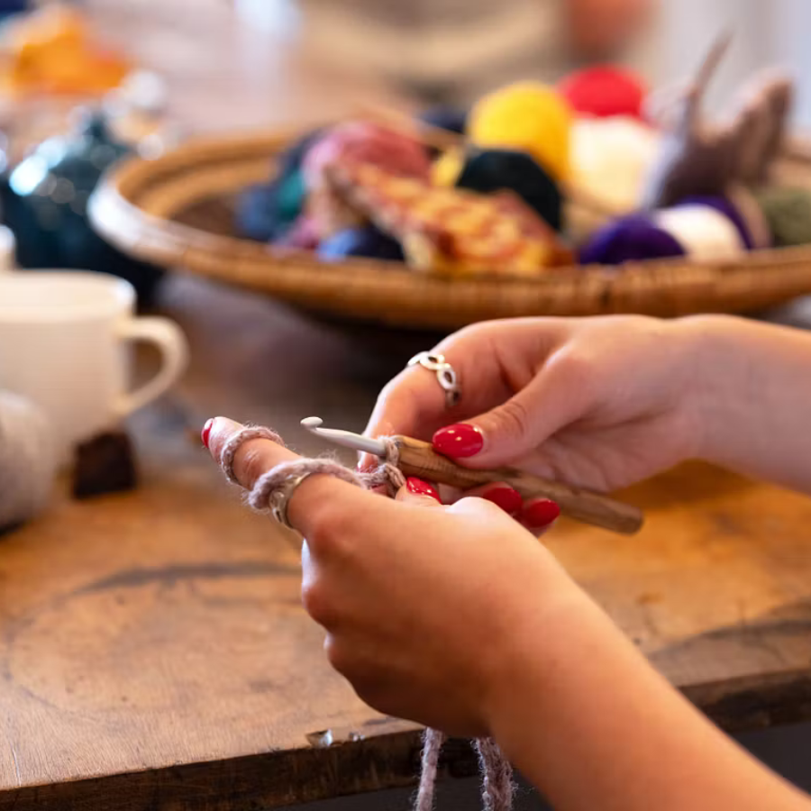 A person with red nail varnish uses a crochet hook and light-coloured yarn at a wooden table. In the background, a tray holds colourful balls of yarn—perfect inspiration for creative London workshops—and there is a white cup nearby.