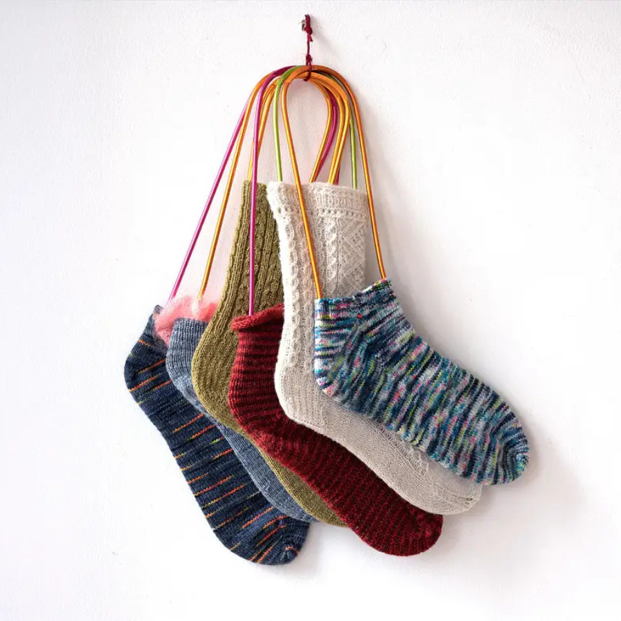 Six colourful knitted socks hang side by side on yellow and pink loops against a white wall, reminiscent of designs crafted in London workshops, showcasing patterns and textures in blue, green, red, white, and multicoloured yarns.