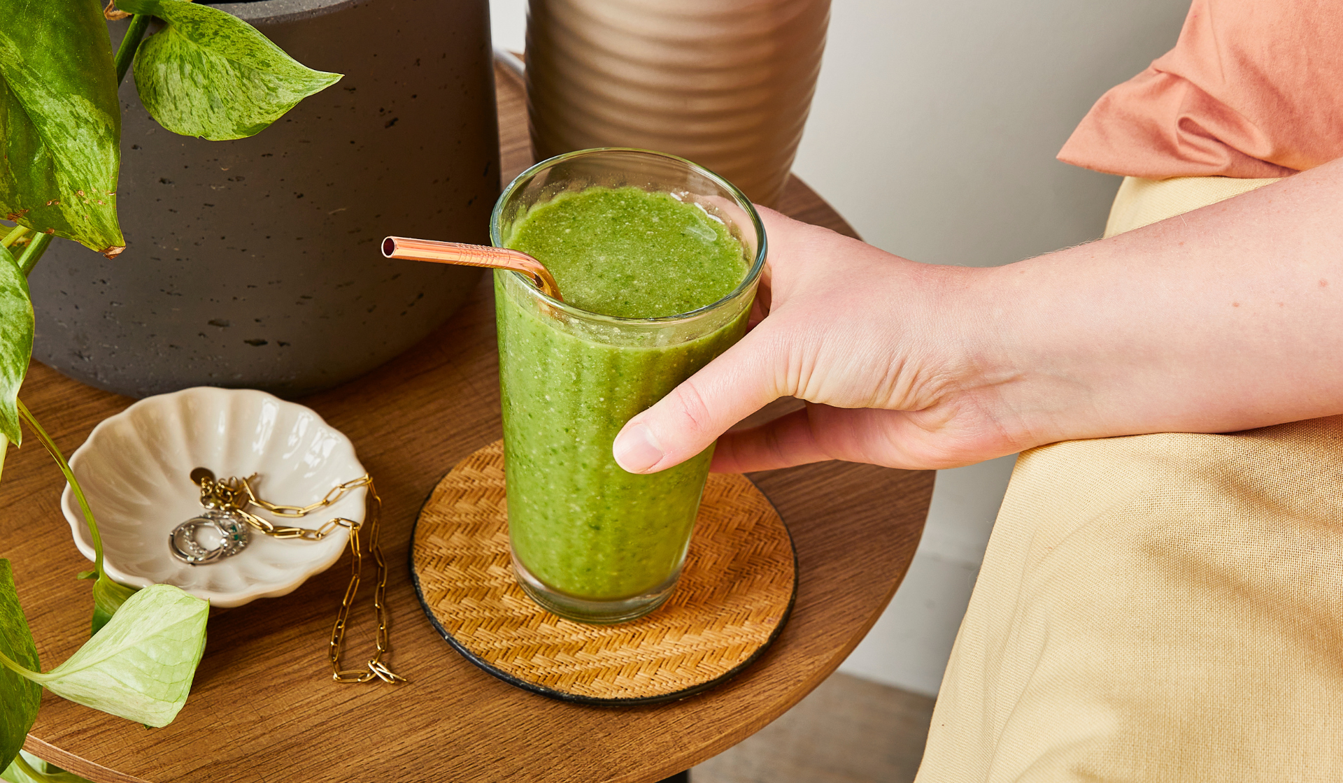 A hand holding a glass of green smoothie with a metal straw, placed on a coaster on a wooden table next to jewellery, a plant, Mindful Chef decor, and other decorative items.