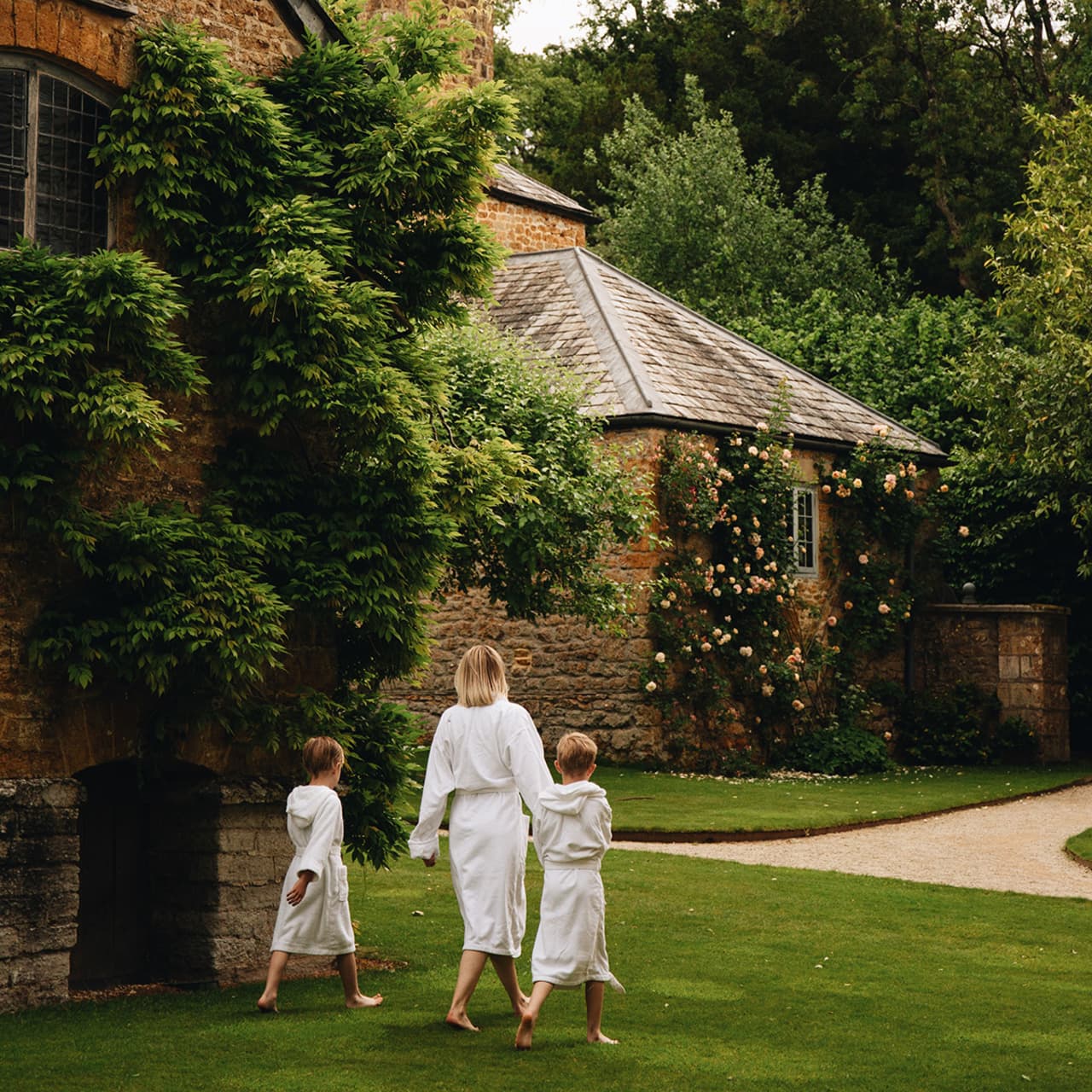 A woman and two children, all in white robes, walk barefoot on a garden path beside a stone house covered in green vines—a tranquil escape reminiscent of the best hotels accessible by a scenic train journey from London.