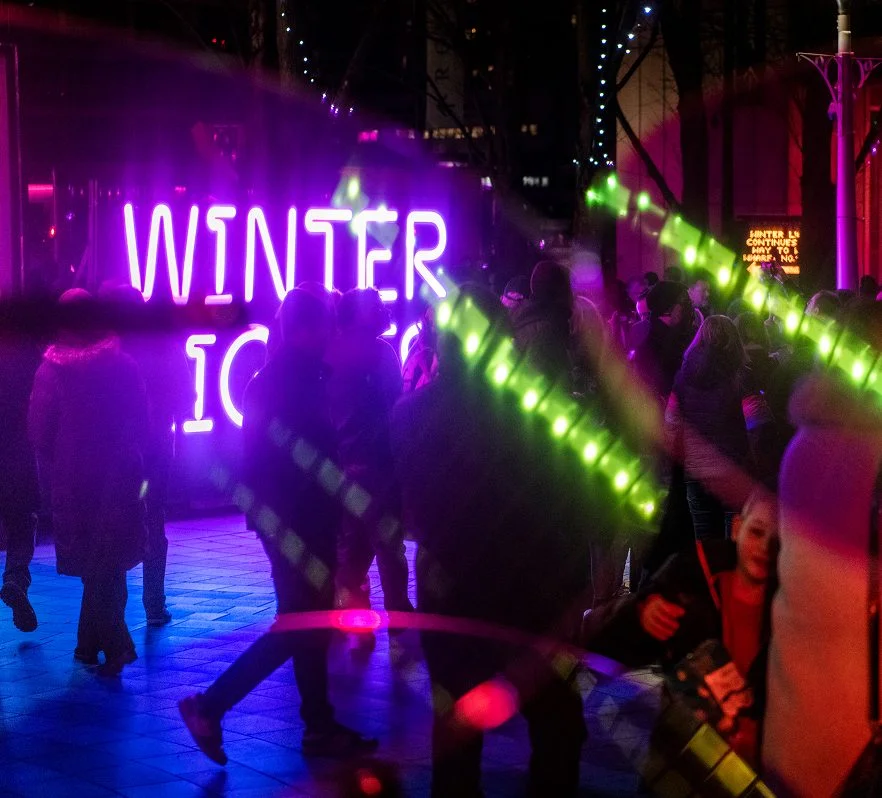 A crowd gathers at night near a bright neon sign that reads WINTER LIGHTS. Green lights and reflections add to the festive atmosphere—an ideal setting for winter date ideas as people wrapped up in cosy clothing enjoy the event.