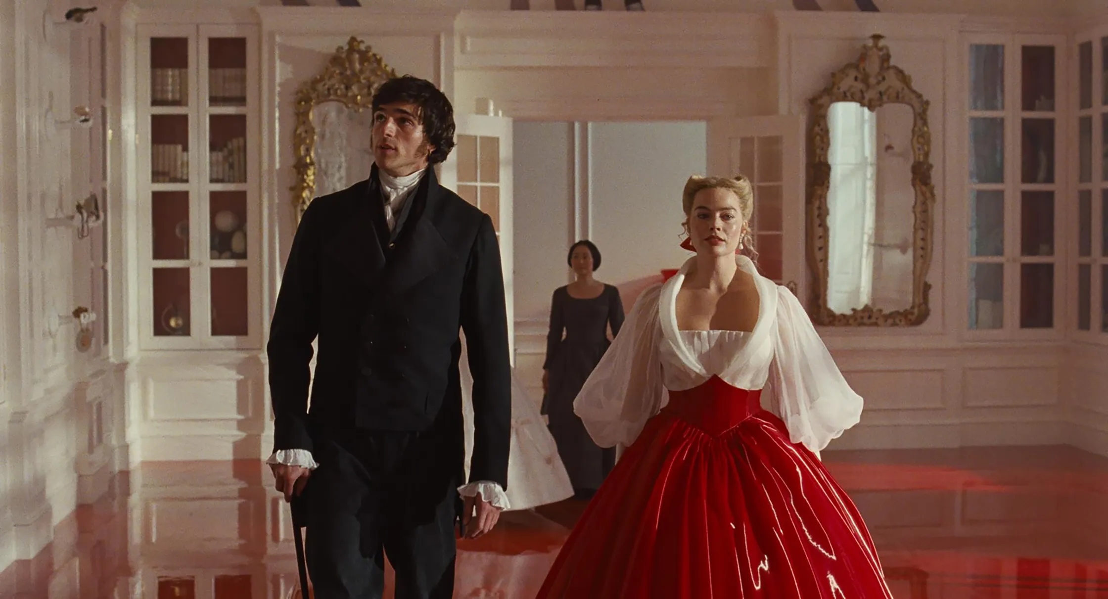A man in a black suit and a woman in a red and white gown walk through an elegant, white room with large mirrors and ornate gold frames—an enchanting scene perfect for winter date ideas—whilst another woman stands in the background.