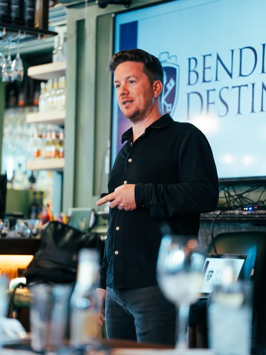 A man in a black shirt stands and speaks in a modern bar or restaurant, giving pub lectures as a screen behind him displays the words BEND and DESTIN. Glasses and blurred objects are visible in the foreground.