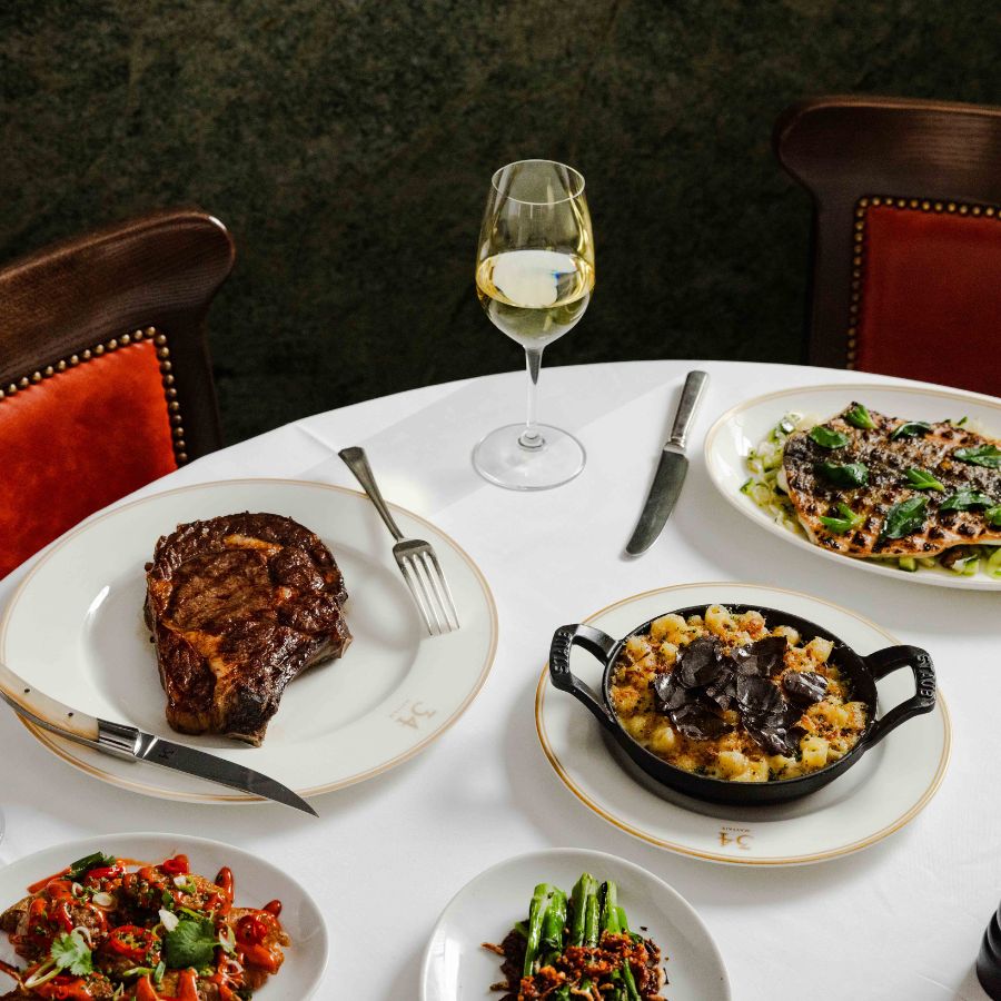 A white tablecloth table set with steak, pasta with truffles, vegetables, and a glass of white wine—perfect for your WEEKEND GUIDE to elegant dining. Two leather chairs are partially visible in the background.