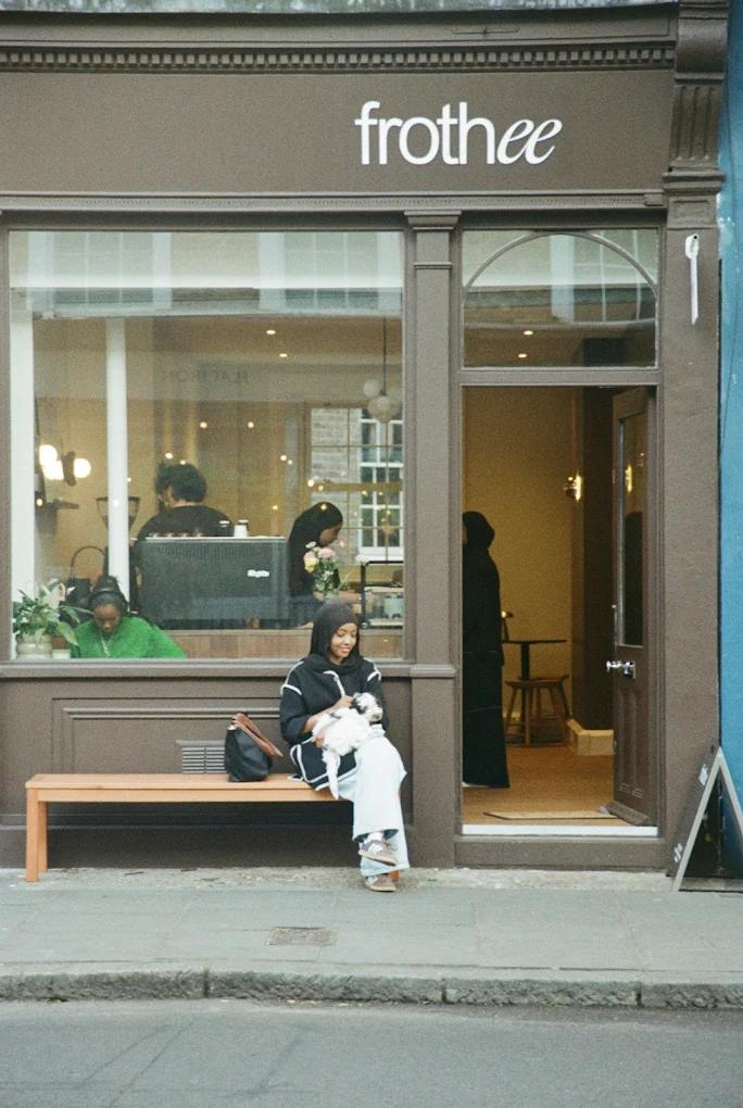 A woman enjoys solo dating on a wooden bench outside a café called frothee, holding a drink and looking at her mobile. The café’s large windows reveal several people inside and the cosy interior.