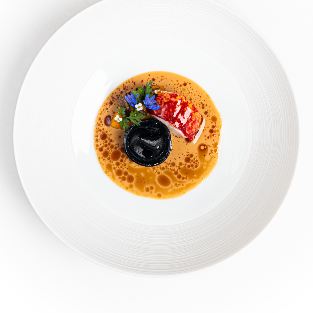 A gourmet dish on a white plate featuring lobster, a black circular element, edible flowers, herbs, and a light brown foam. Artistic and minimalist, this creation is part of an affordable tasting menu that showcases exceptional flavours.