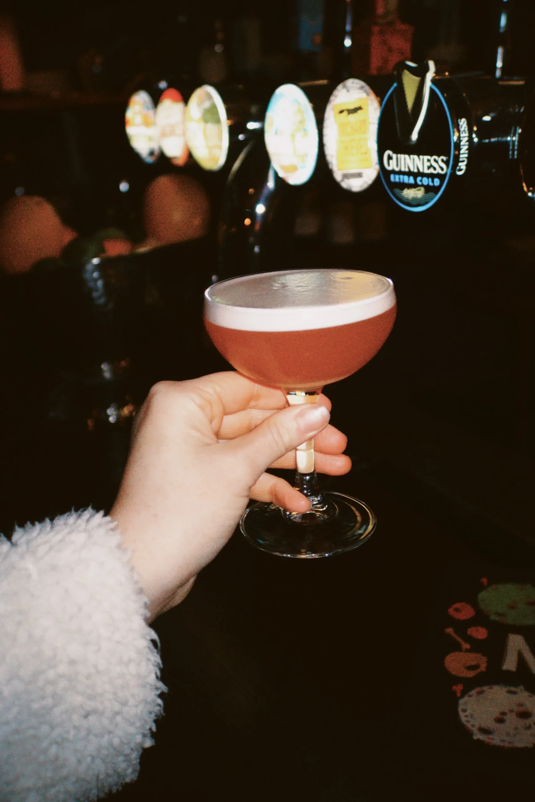 A hand in a fluffy white sleeve holds a coupe glass with a pink cocktail and frothy top at the bar, where beer taps and drinks labels line the backdrop—capturing the vibrant spirit of the margarita mile.