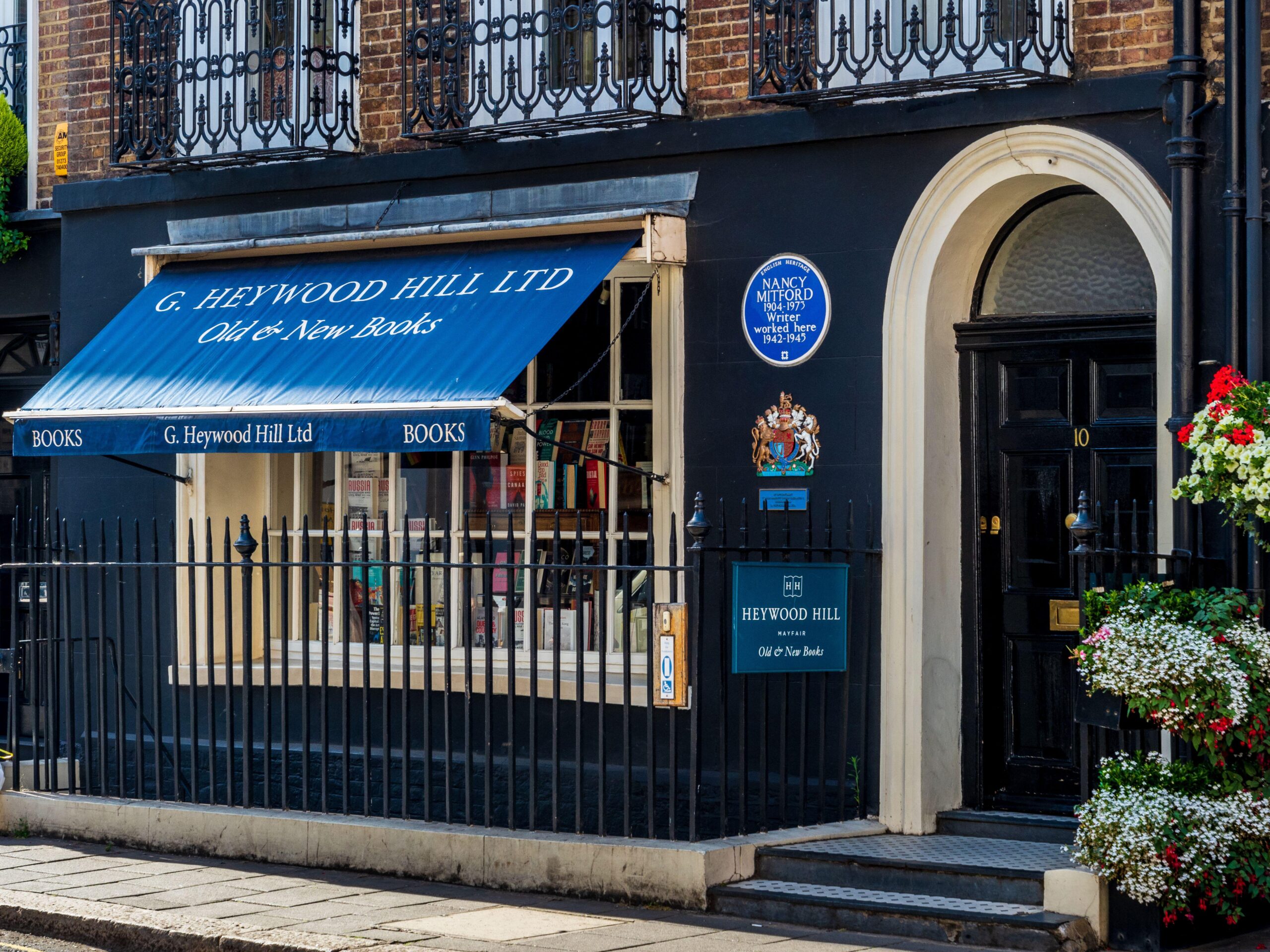 A bookshop with a blue awning reading G. Heywood Hill Ltd Old & New Books and a window displaying books. One of the best London bookshops, it features a blue plaque above and flowers hanging near the entrance at 10 Curzon Street.