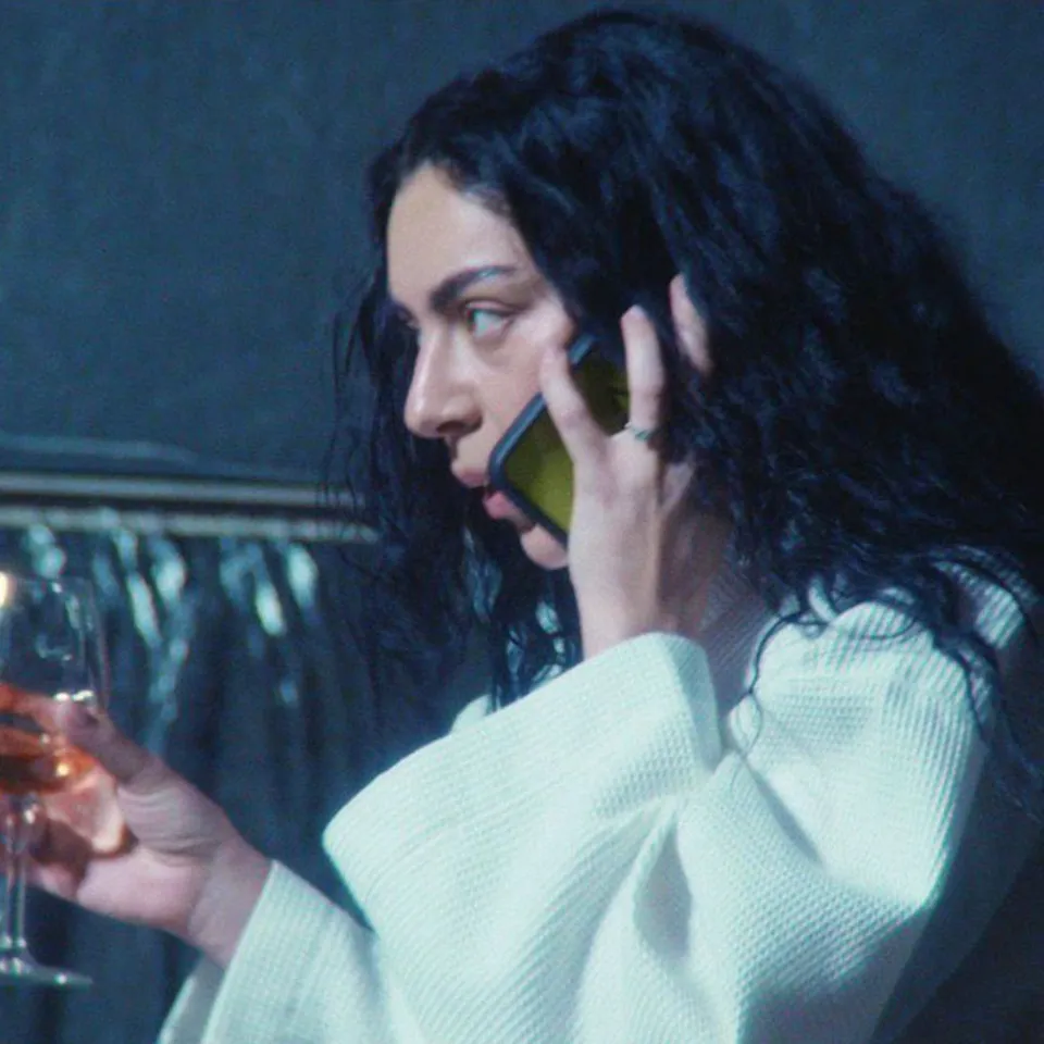 A person with long, dark hair wearing a white dressing gown holds a wine glass in one hand and talks on a green phone with the other, looking to the side—evoking the stylish vibe of Charli XCX in a new film scene.