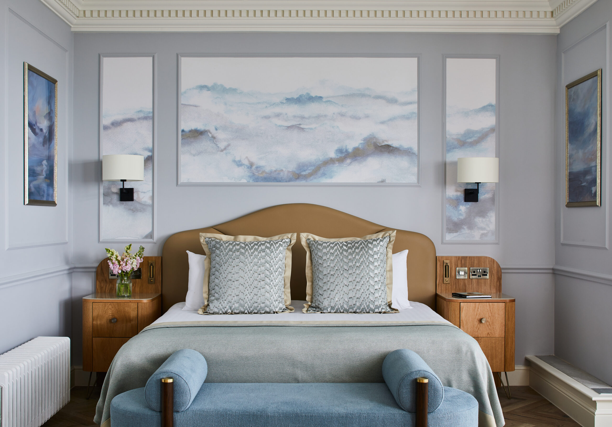 A stylish, modern bedroom reminiscent of those in Brighton hotels, featuring a large bed with a beige headboard, blue bench, pillows, and soft blue-green bedspread. Abstract artwork hangs above the bed, flanked by matching bedside tables and lamps.