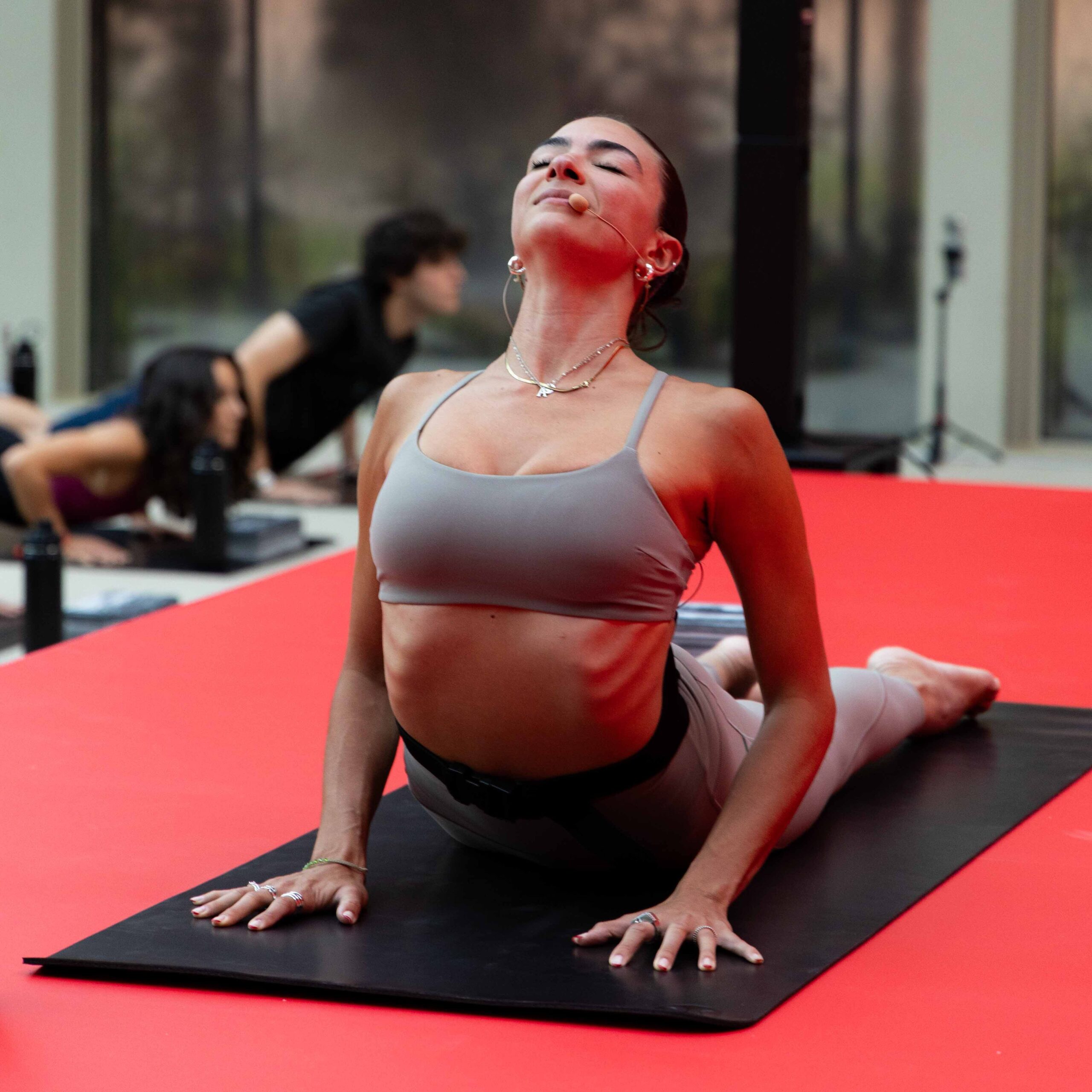 A woman in sportswear performs an upward-facing dog yoga pose on a black mat atop a red floor, with other people practising yoga in the background—perfect inspiration for your weekend guide.